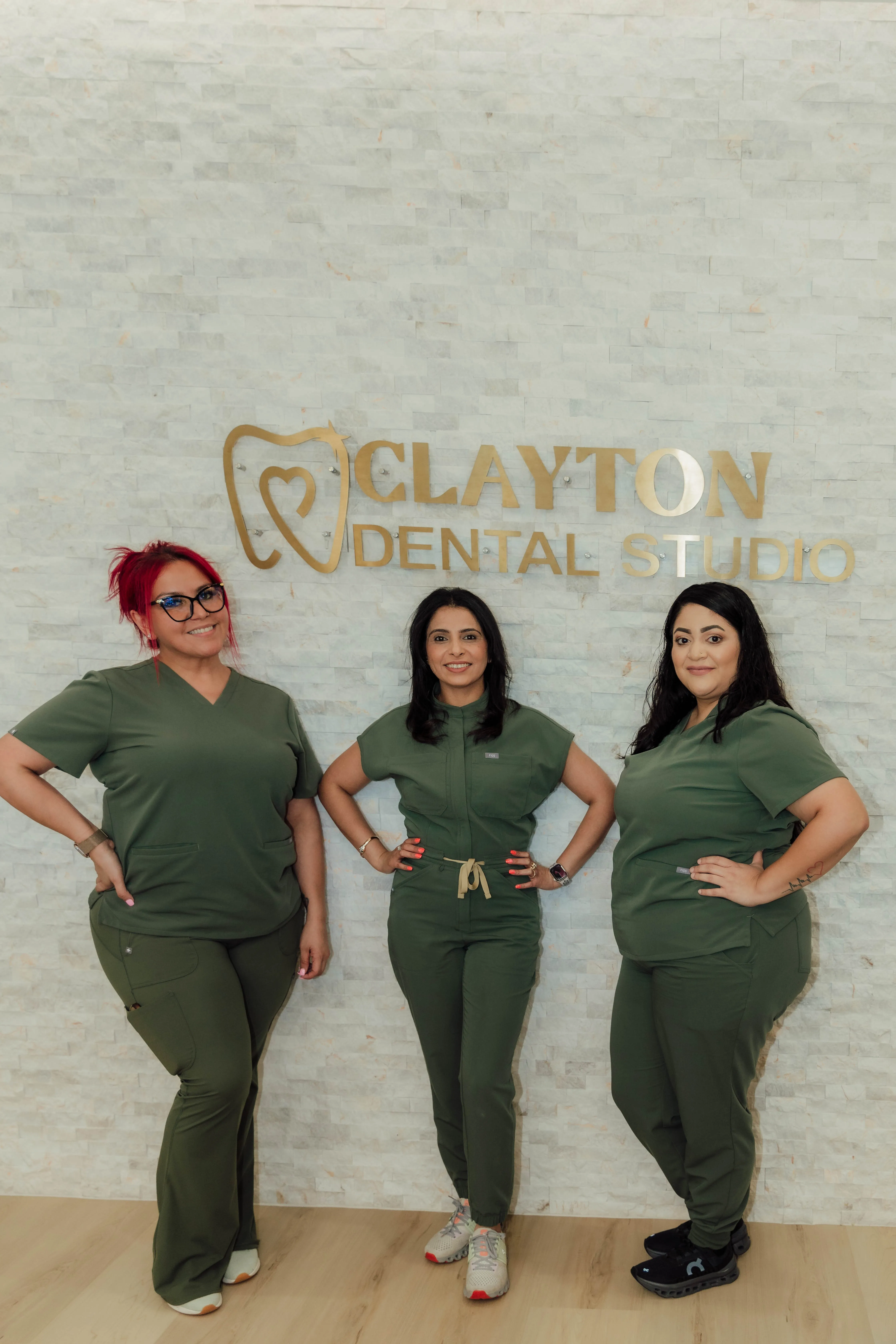 Three women in green medical scrubs standing and smiling in front of a light-colored wall with a gold Clayton Dental Studio logo.