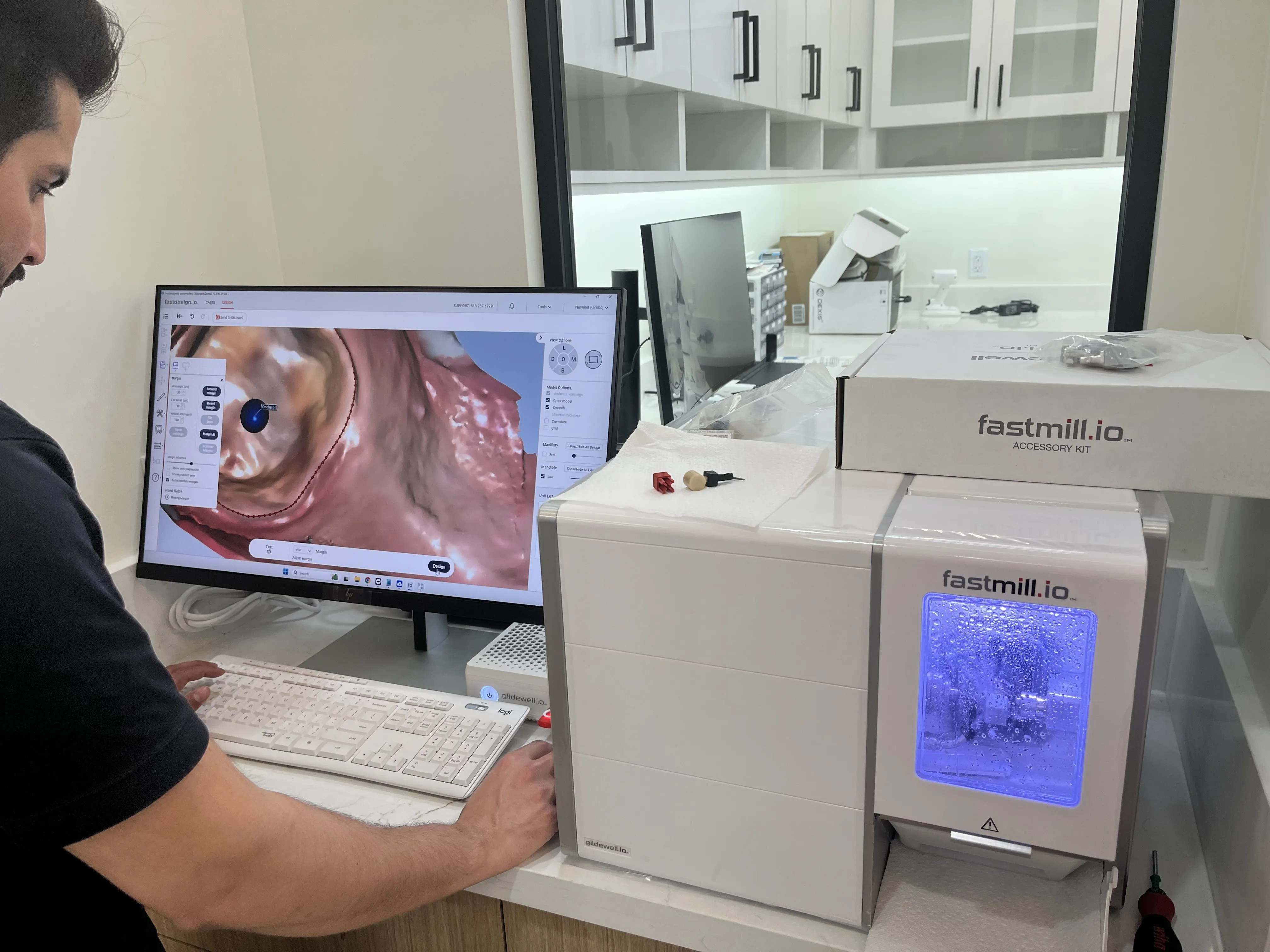 Person working on a dental design software displayed on a monitor next to a white fastmill.io milling machine with a glowing blue window.