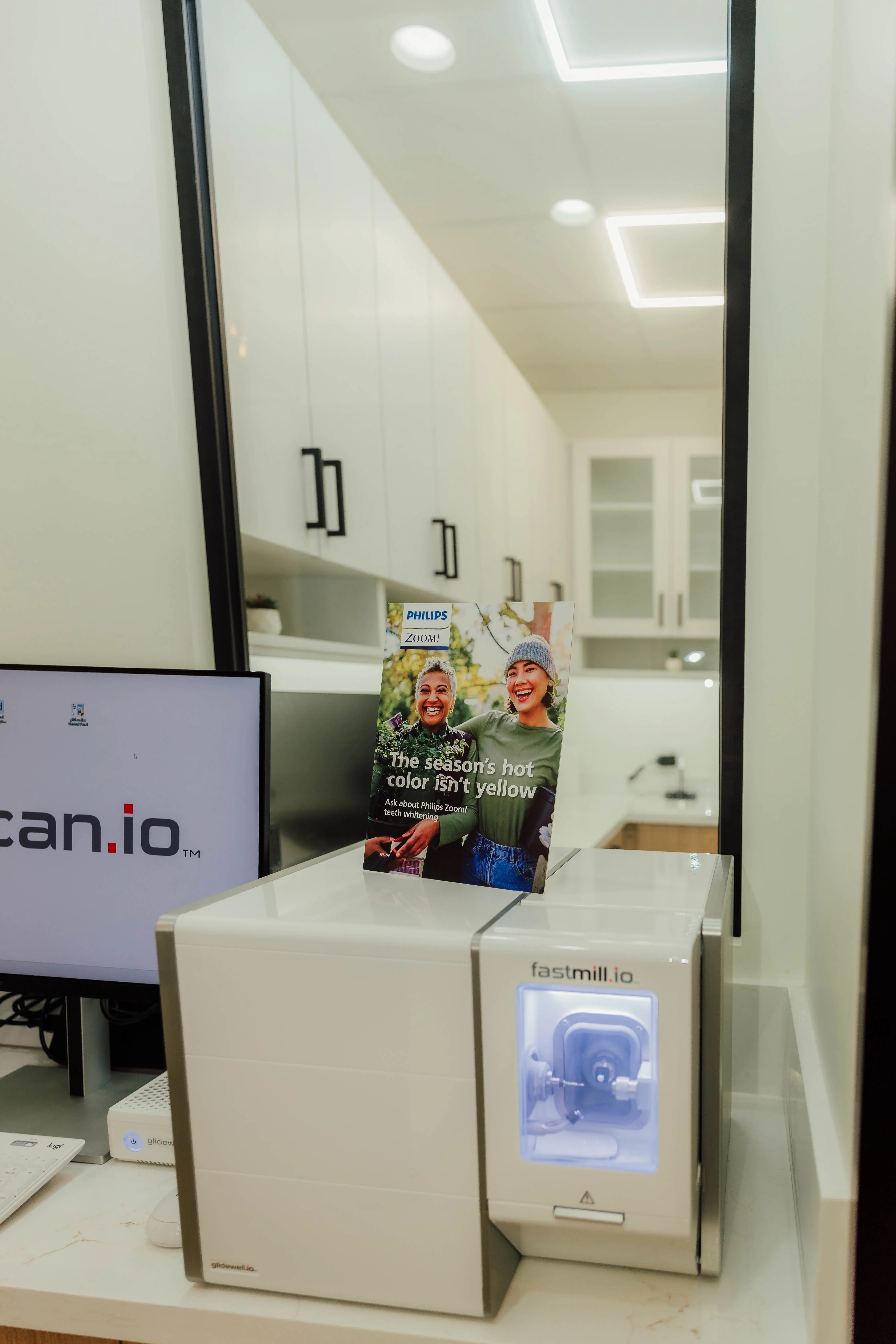 Fastmill.io dental milling machine on a countertop with a Philips Zoom teeth whitening leaflet and a computer monitor in a dental office.