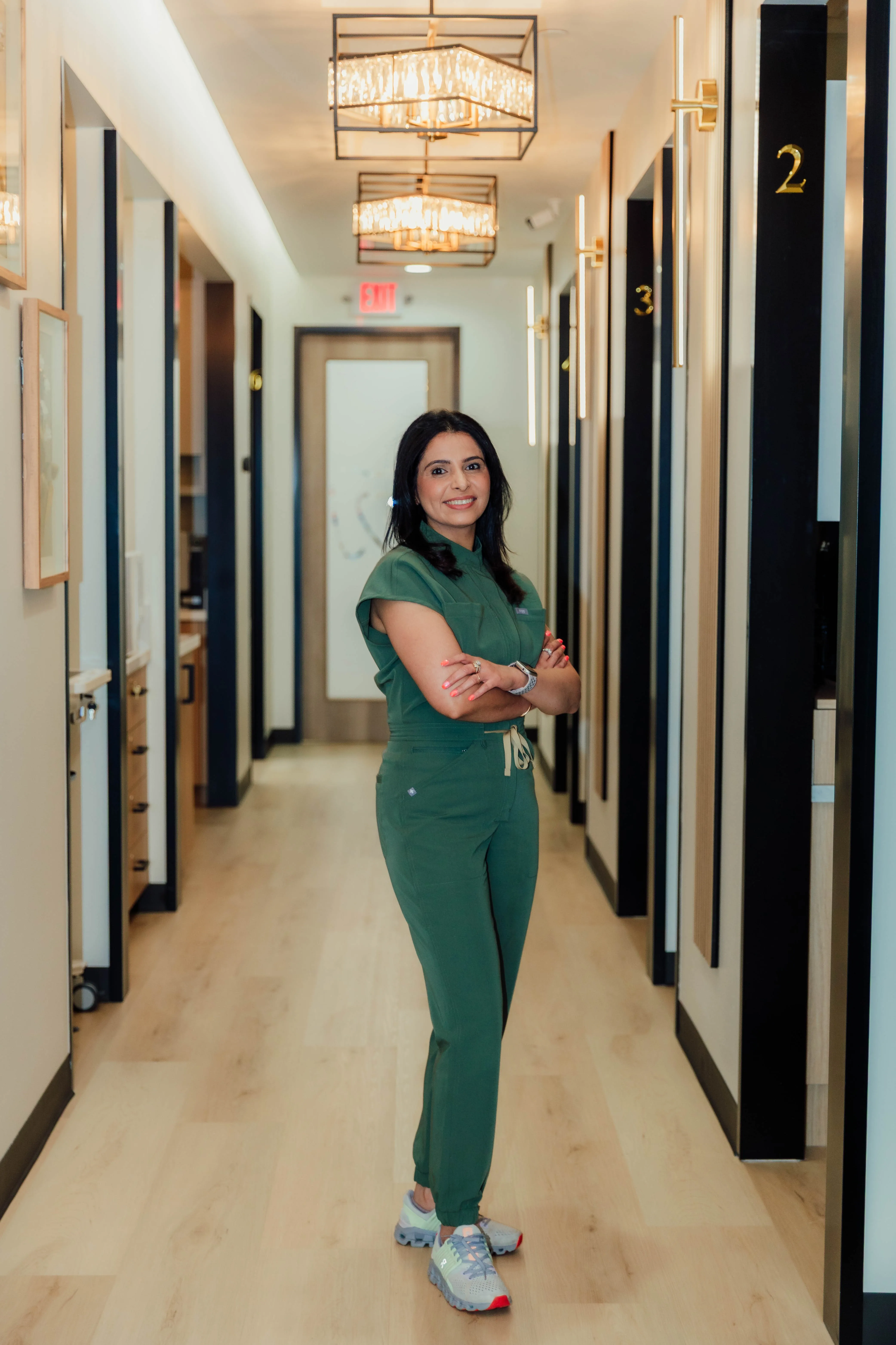 Smiling woman in green medical scrubs standing with arms crossed in a brightly lit hallway with numbered doors and wooden floors.