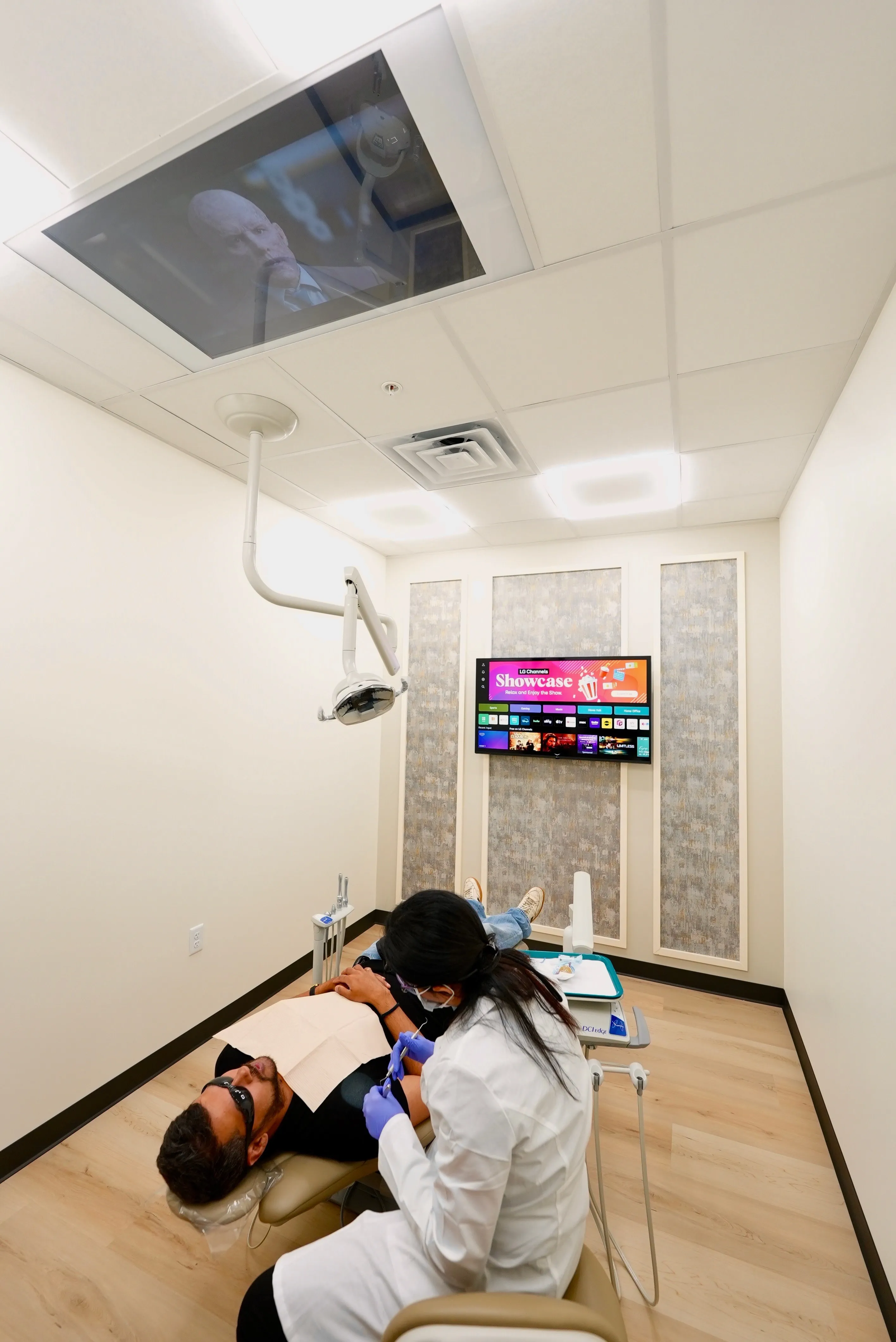 Dentist wearing gloves and mask treating a patient reclining in a dental chair with a TV screen on the wall and a ceiling-mounted screen showing a movie scene.
