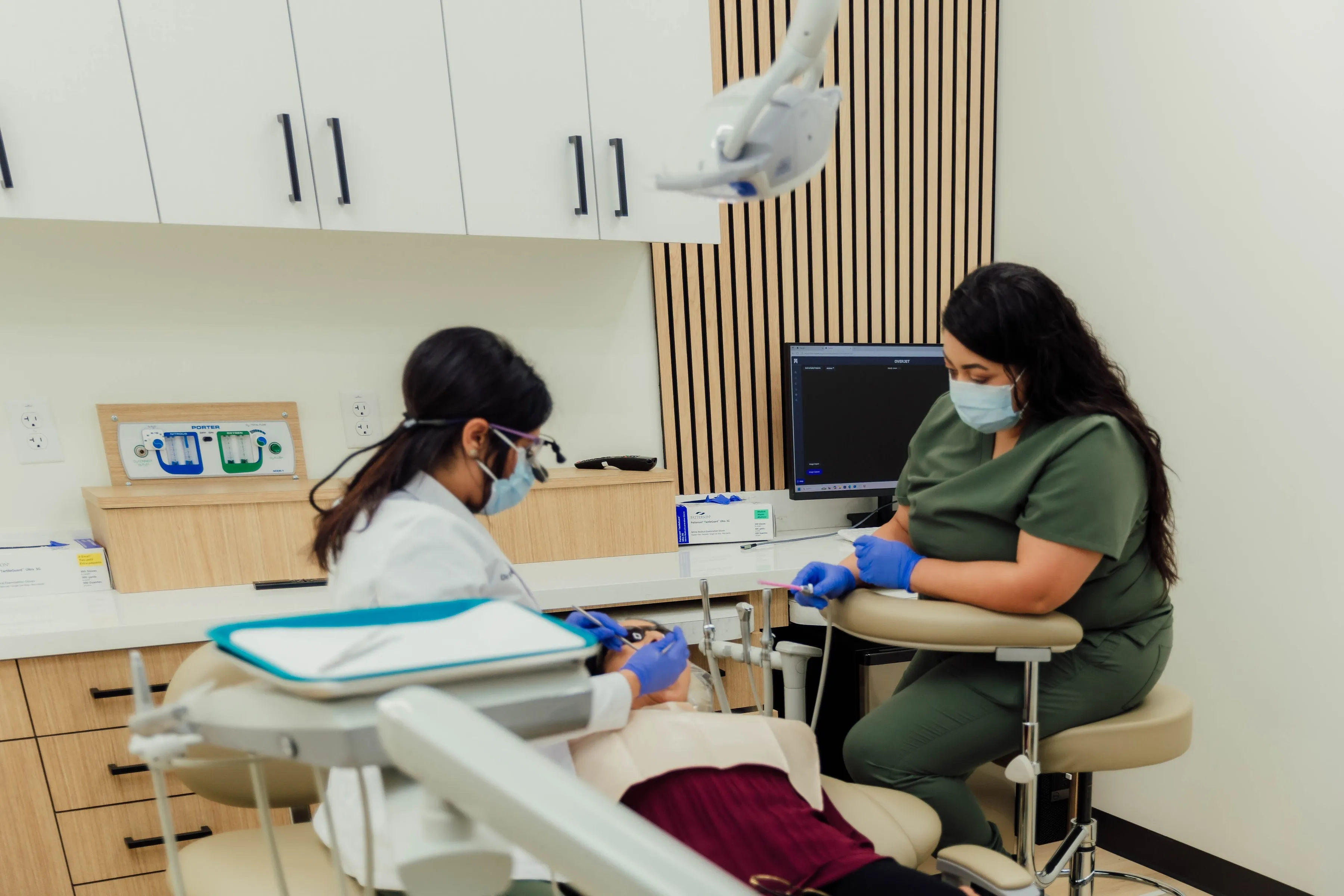 A female dentist Dr. Dr. Navneet Kamboj and an assistant wearing masks and gloves attending to a patient lying on a dental chair in a modern clinic.