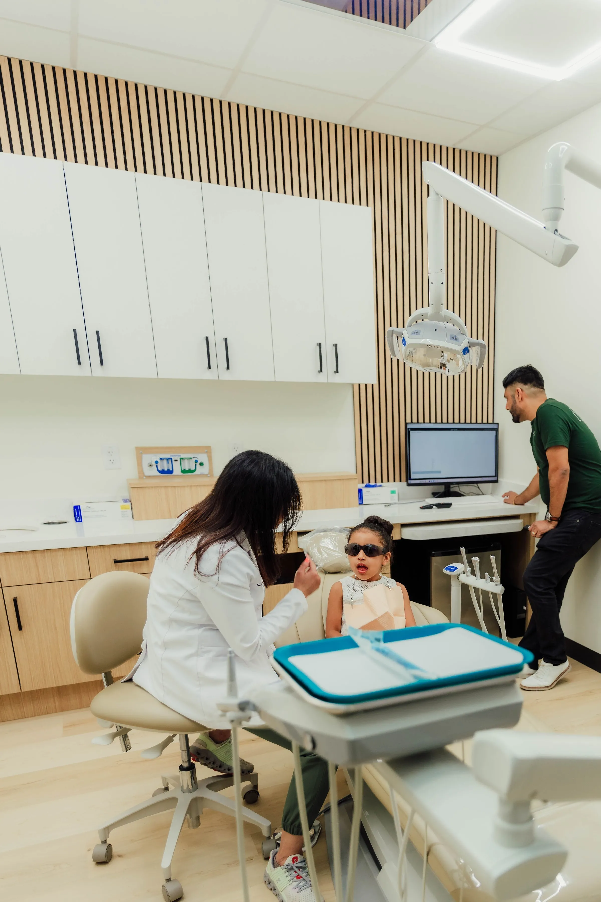 Dentist talking to a young child wearing sunglasses seated in a dental chair, with a man observing near a computer monitor in a modern dental office.