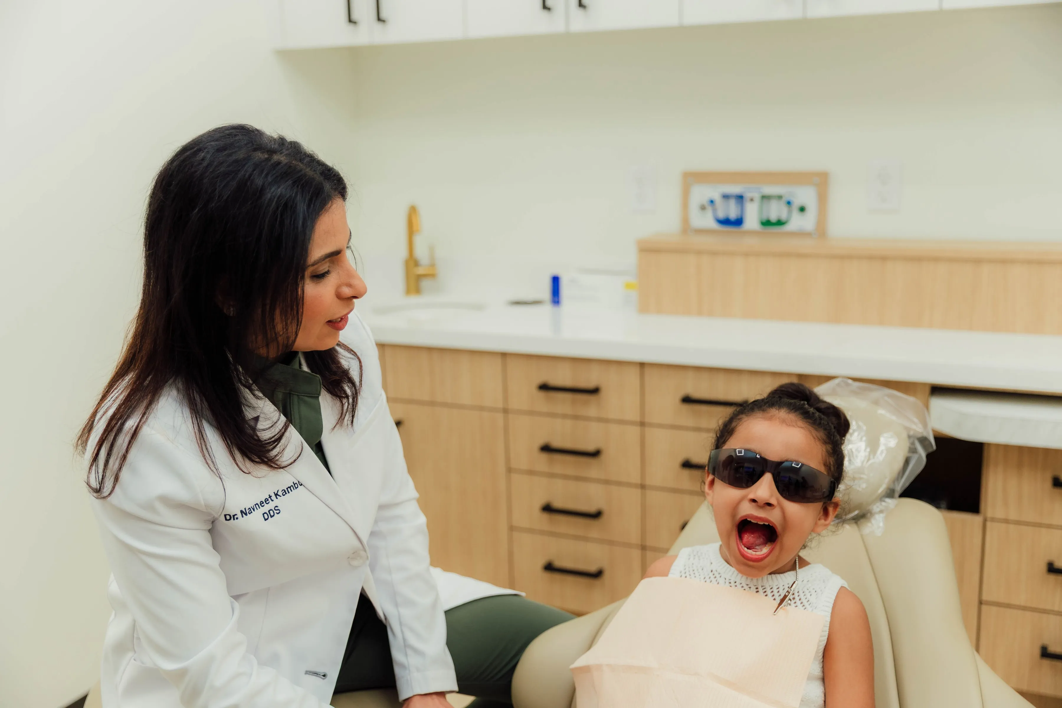 Child wearing dark sunglasses sitting in a dental chair with mouth open, a female dentist in a white coat talking to her.