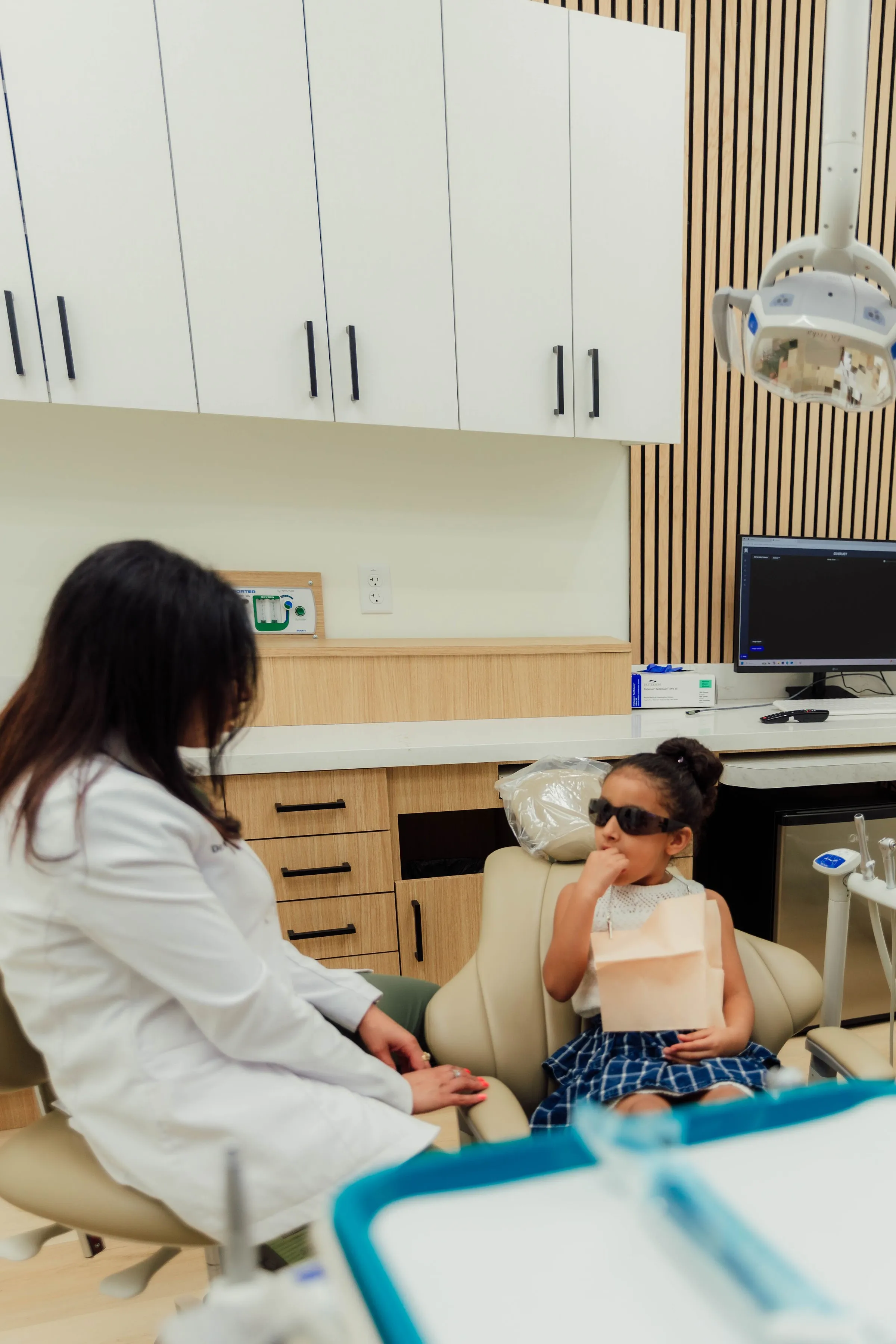 Young girl wearing sunglasses sitting in a dental chair with a bib, while a female dentist in a white coat talks to her in a modern dental office.