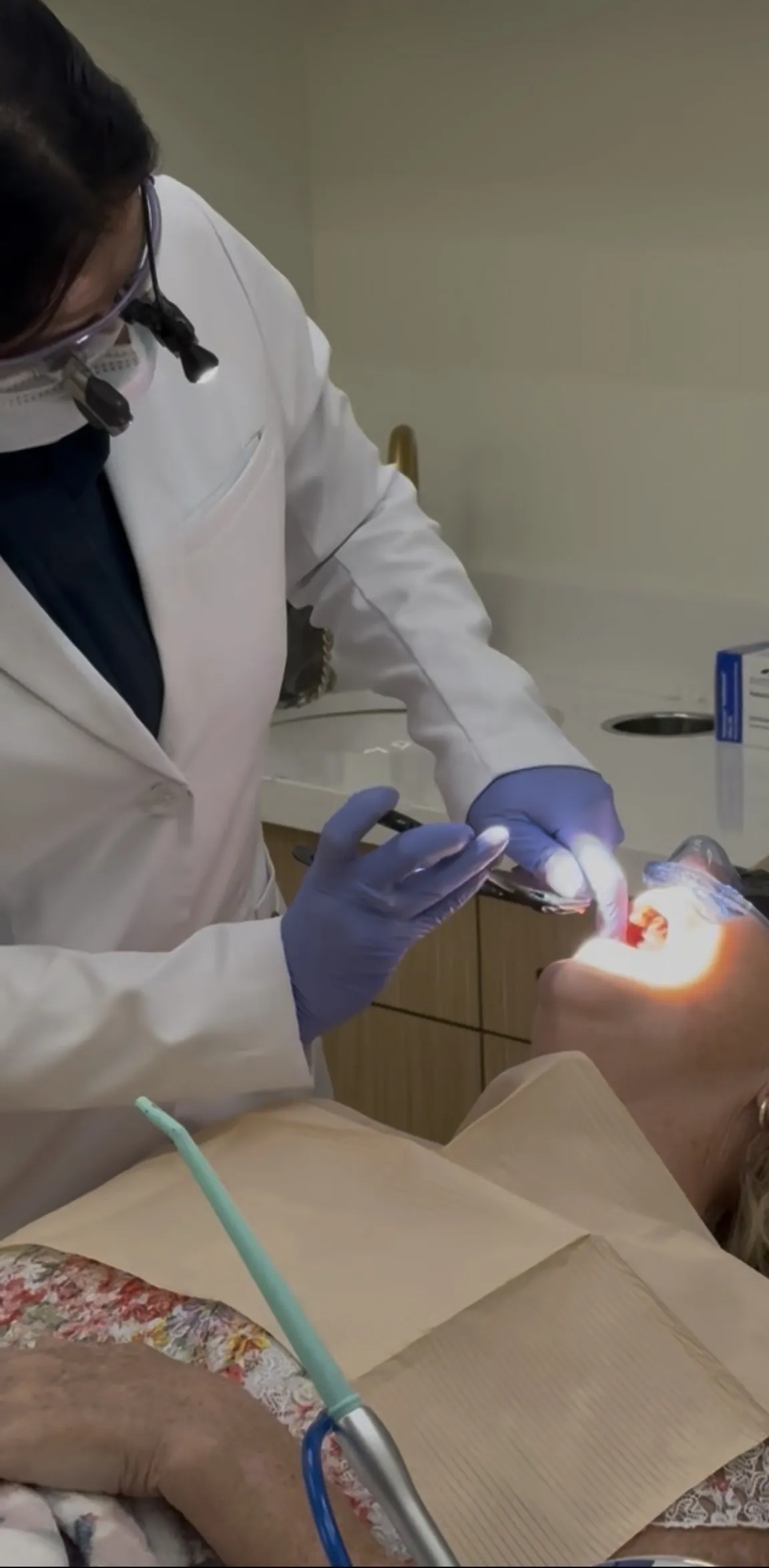 Dentist wearing magnifying glasses and gloves examining a patient's mouth under bright light.