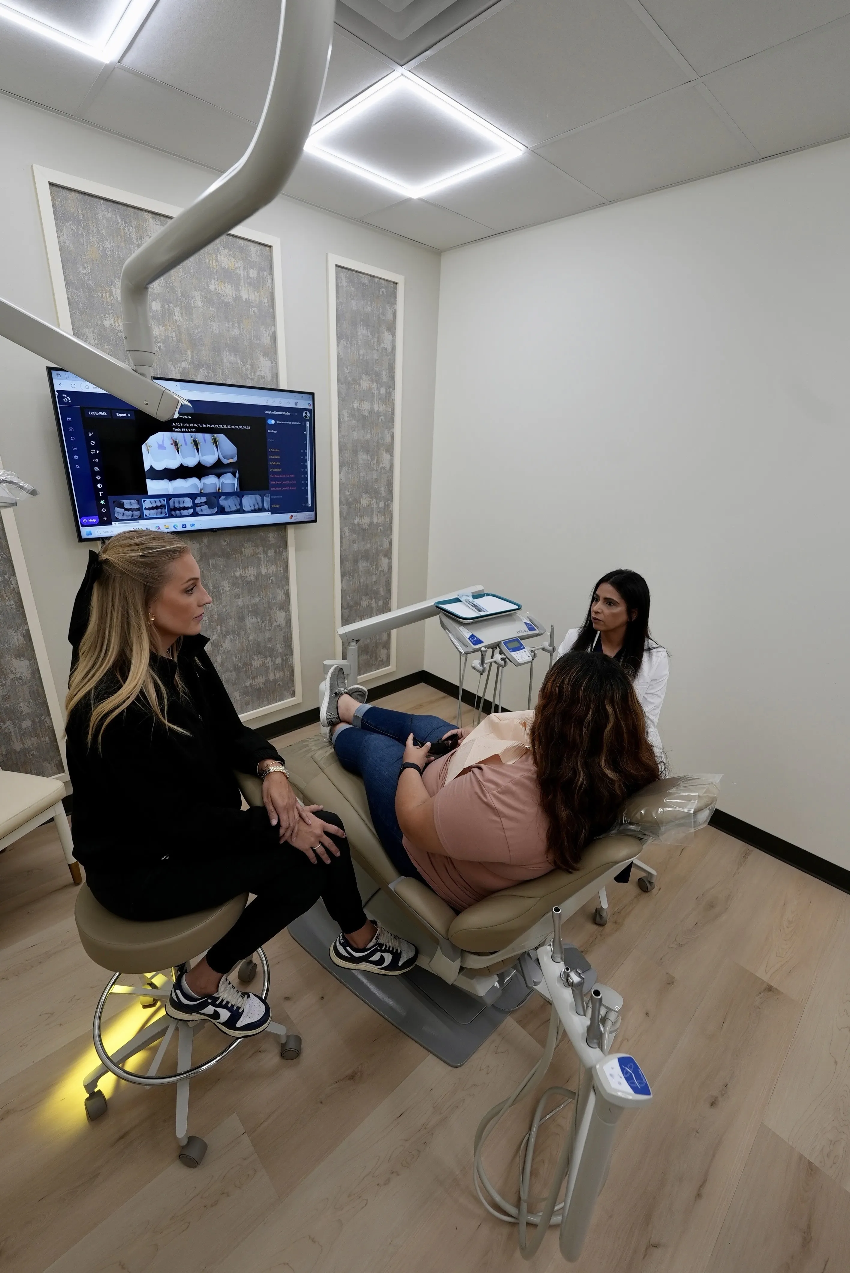 clayton dental studio Dentist and assistant consulting a patient seated in a dental chair with dental x-rays displayed on a screen in the background.