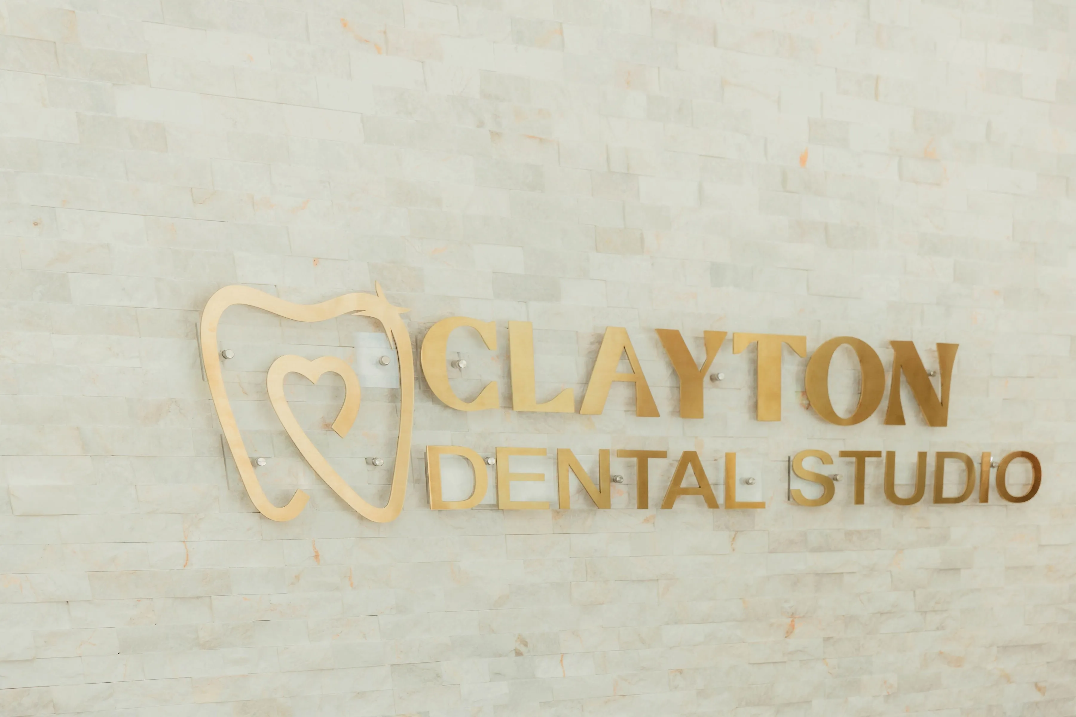Gold metal sign on light stone wall reading Clayton Dental Studio with an abstract tooth and heart logo.