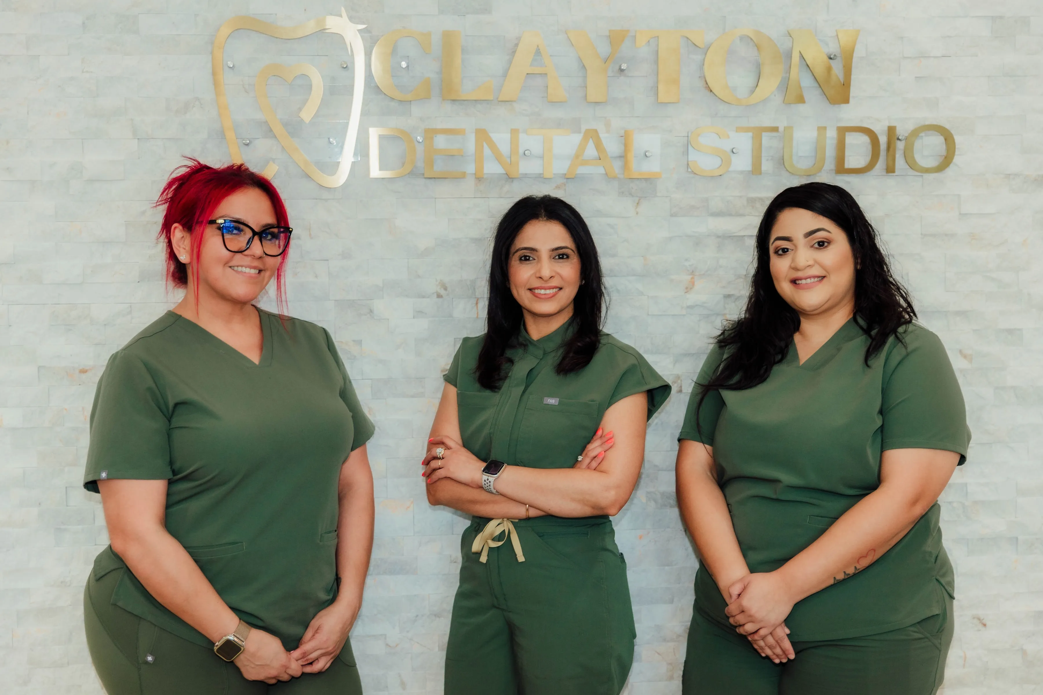 Three women in green scrubs standing and smiling in front of a wall with the gold sign 'Clayton Dental Studio'.