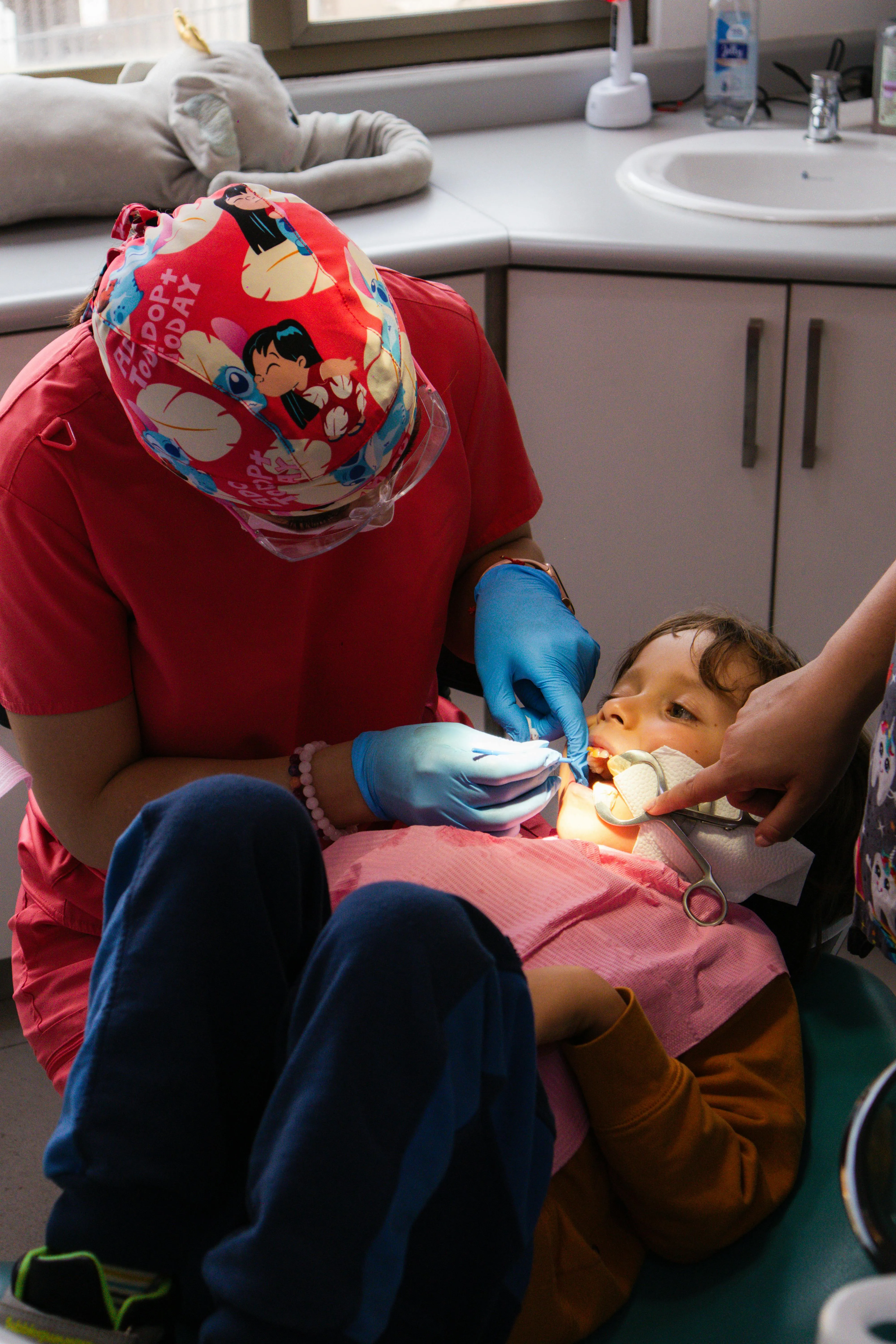Dentist wearing cartoon-themed cap and gloves examining a young boy's teeth in a dental clinic.