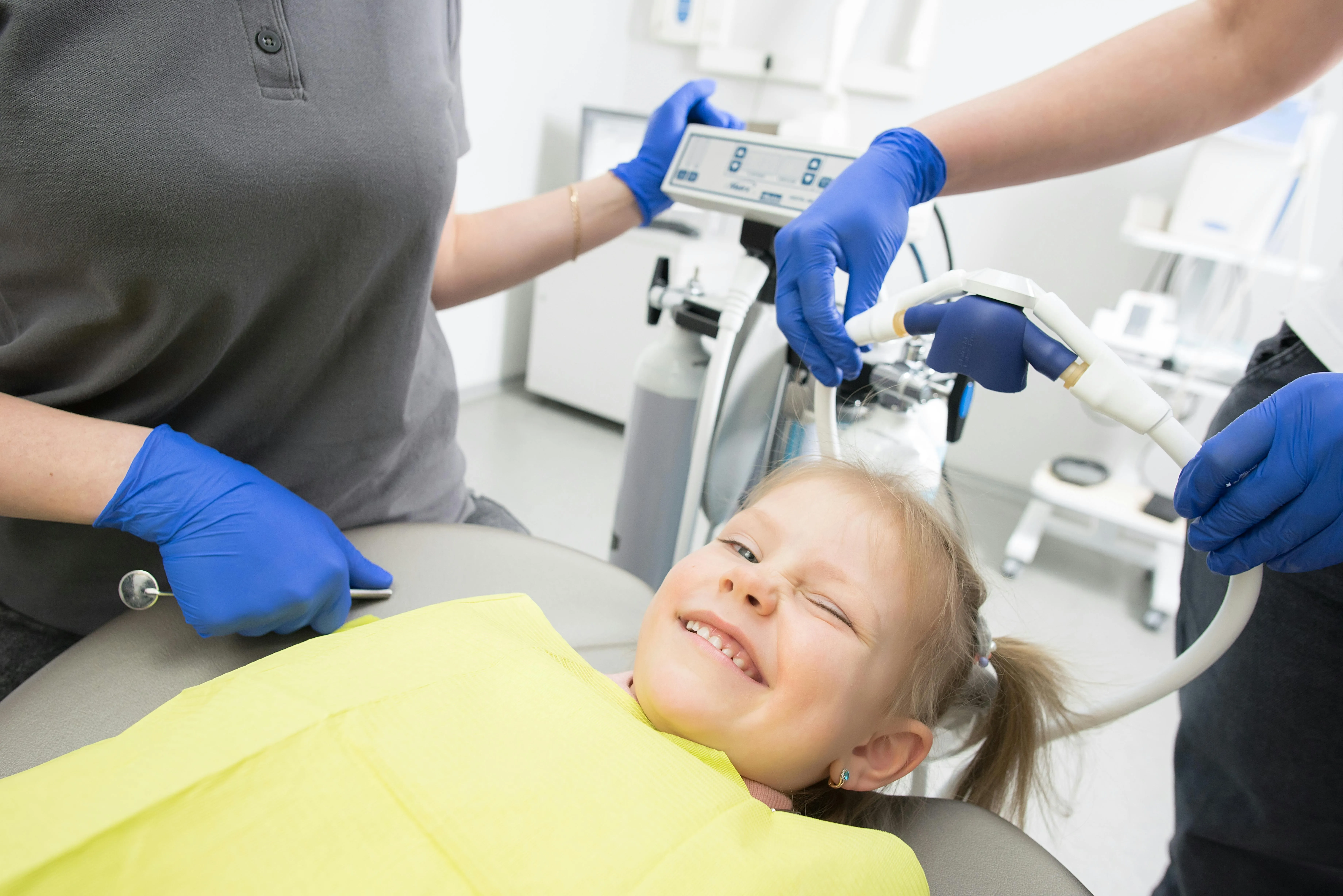 Smiling young girl lying in a dental chair, winking, with two dental professionals wearing blue gloves preparing dental equipment nearby.