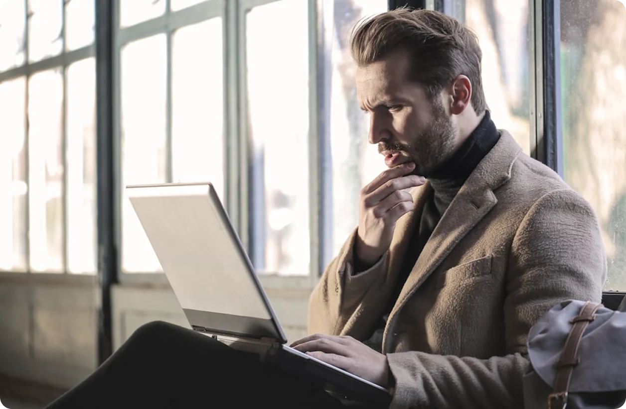 Hombre pensativo usando laptop sentado cerca de una ventana grande.