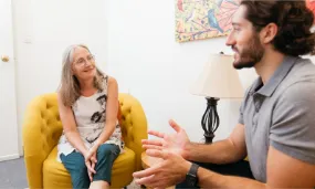 A woman sitting on a yellow armchair attentively listening to a man speaking to her in a cozy room.