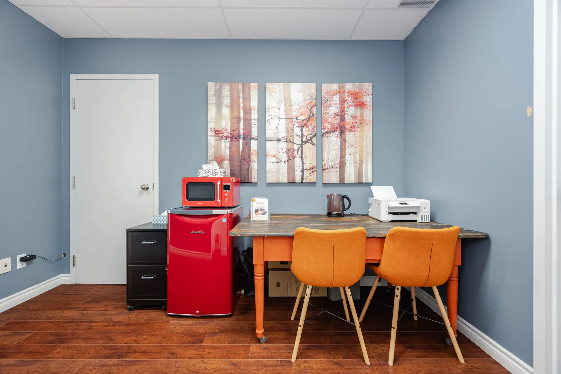 Small office kitchenette with blue walls, a wooden table with two orange chairs, a red mini fridge topped with a red microwave, a printer, and a wall triptych of autumn trees.