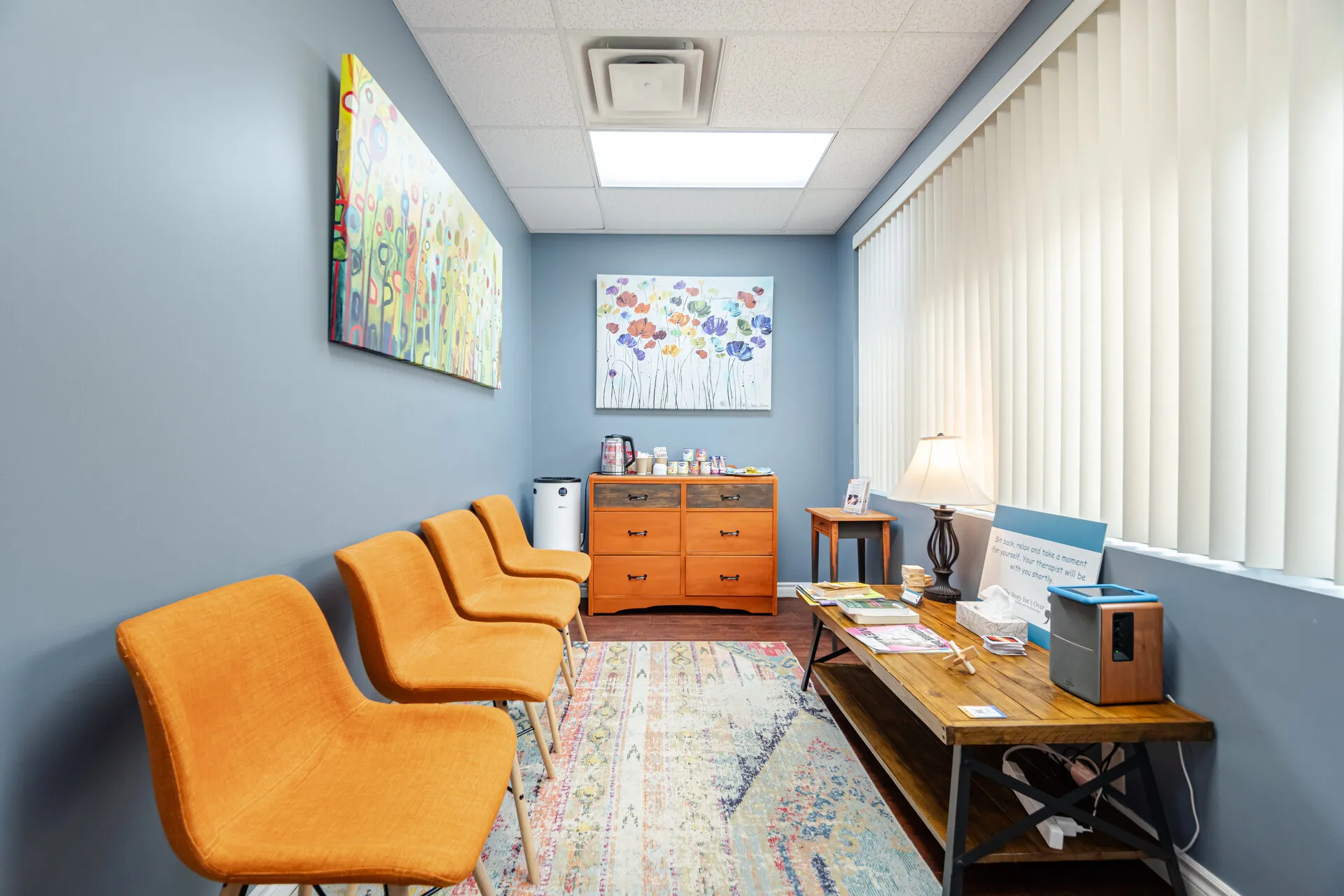 Waiting room with four orange chairs along a blue wall, colorful floral paintings, a wooden cabinet, and a wooden table with magazines and a lamp.