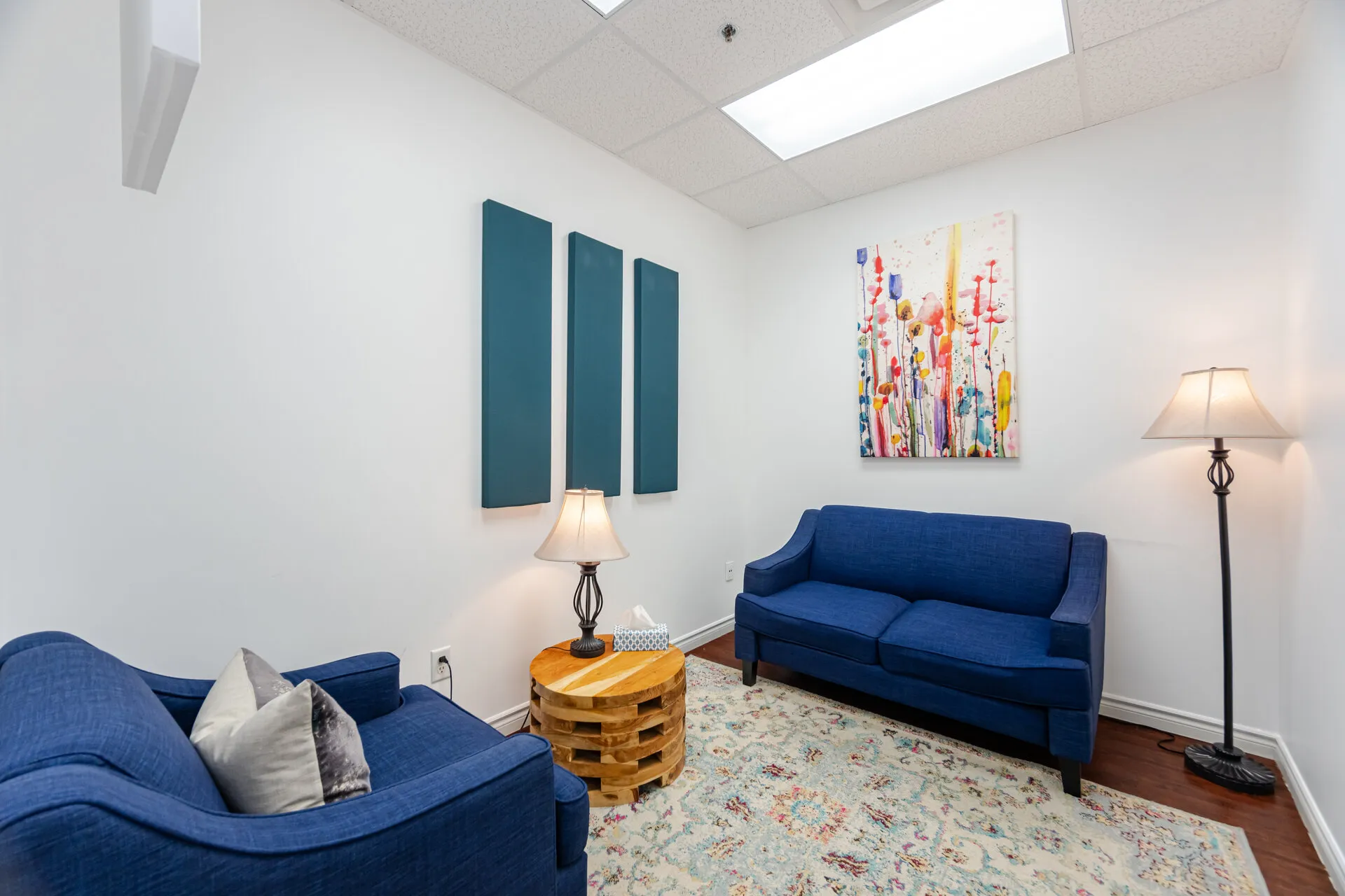 Compact modern sitting area with blue sofa and armchair, wooden side table with lamp, colorful wall art, and patterned rug.