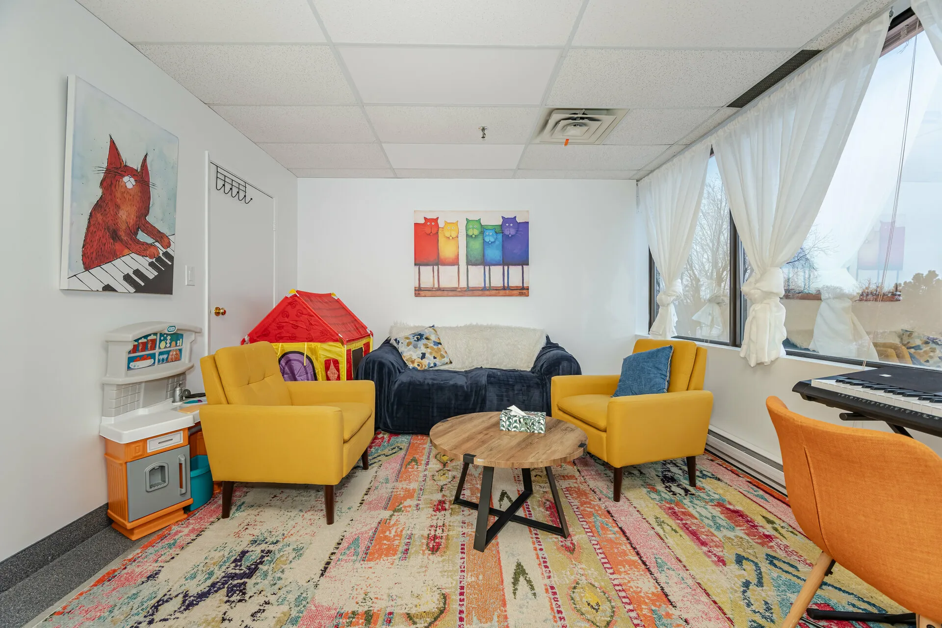 Bright living room with two yellow armchairs, a dark blue sofa, colorful rug, round wooden coffee table, children's play kitchen and playhouse, and cat-themed artwork on the walls.
