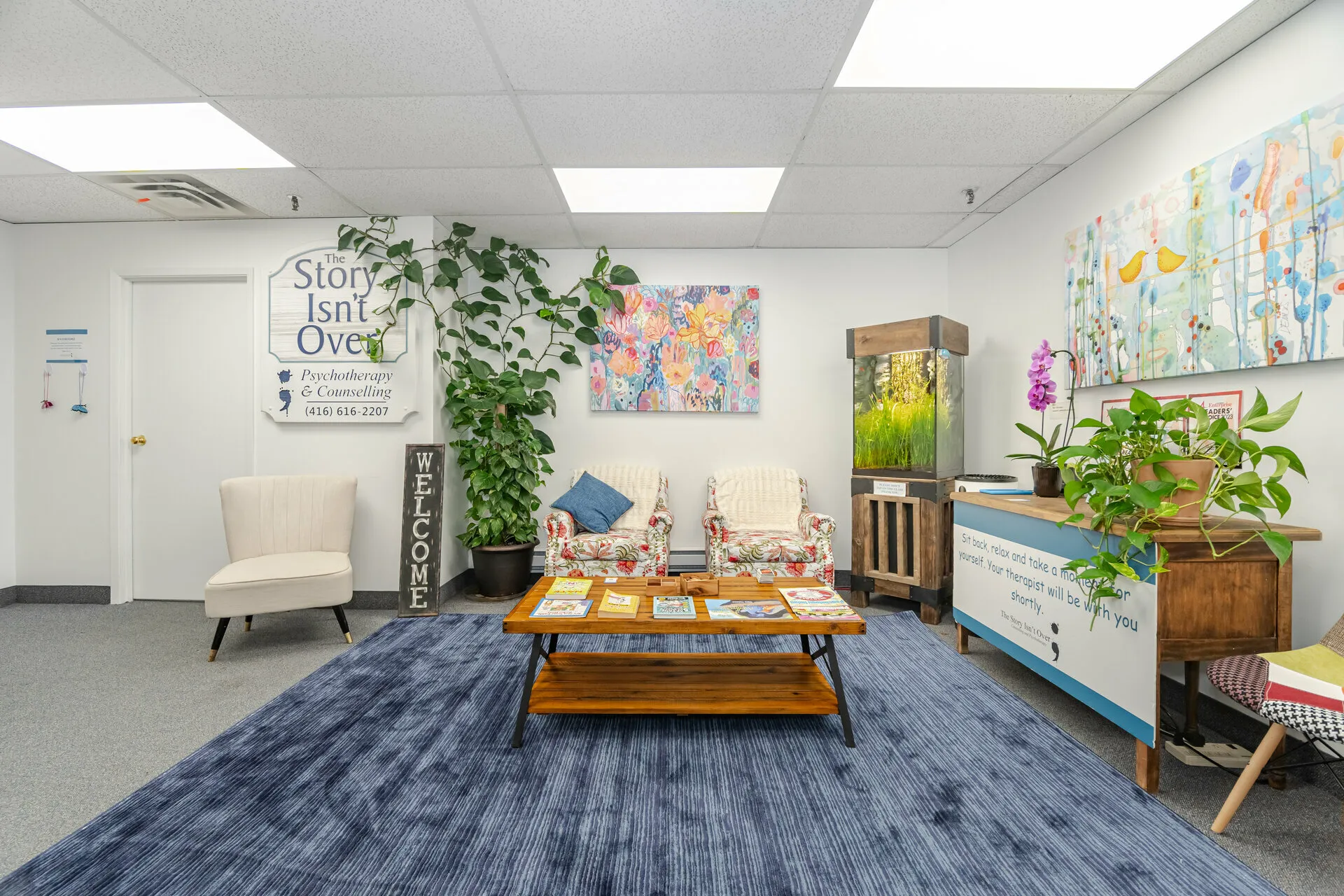 Waiting area with floral armchairs, a wooden coffee table with books, a blue rug, potted plants, fish tank, and colorful wall art in a psychotherapy office.