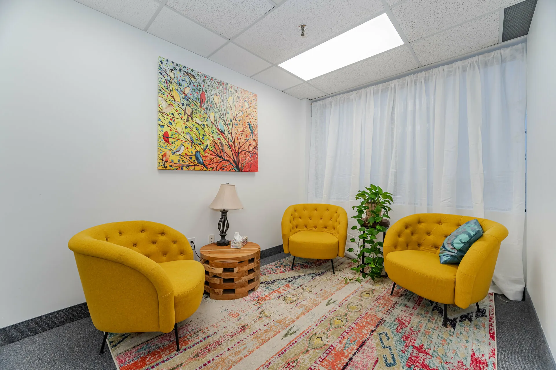Modern seating area with three mustard yellow tufted chairs, a round wooden side table with a lamp and tissues, a green potted plant, a colorful patterned rug, and a bird-themed painting on the wall.