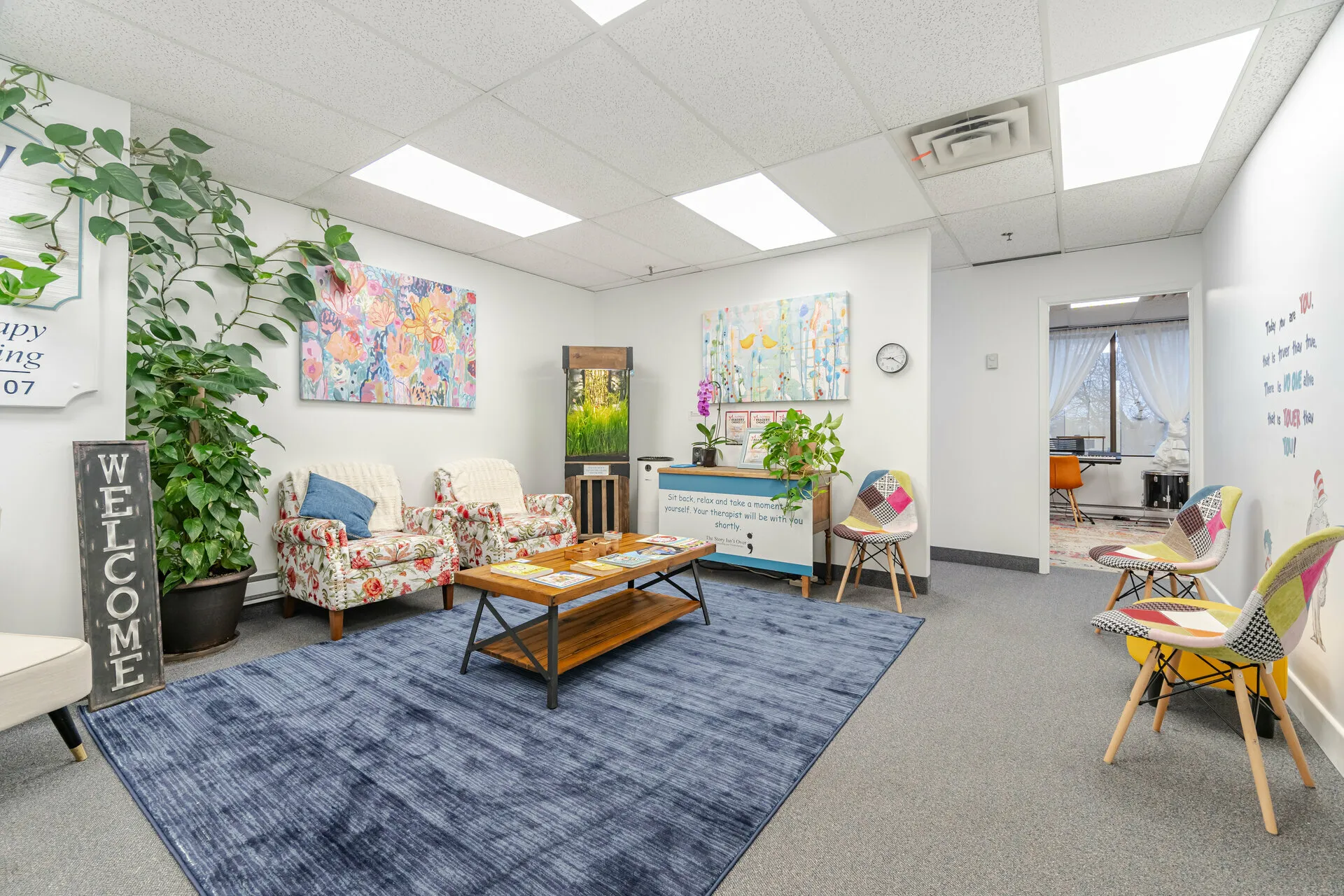 Bright waiting room with floral armchairs, colorful patchwork chairs, a blue rug, plants, artwork on white walls, and a wooden coffee table with magazines.