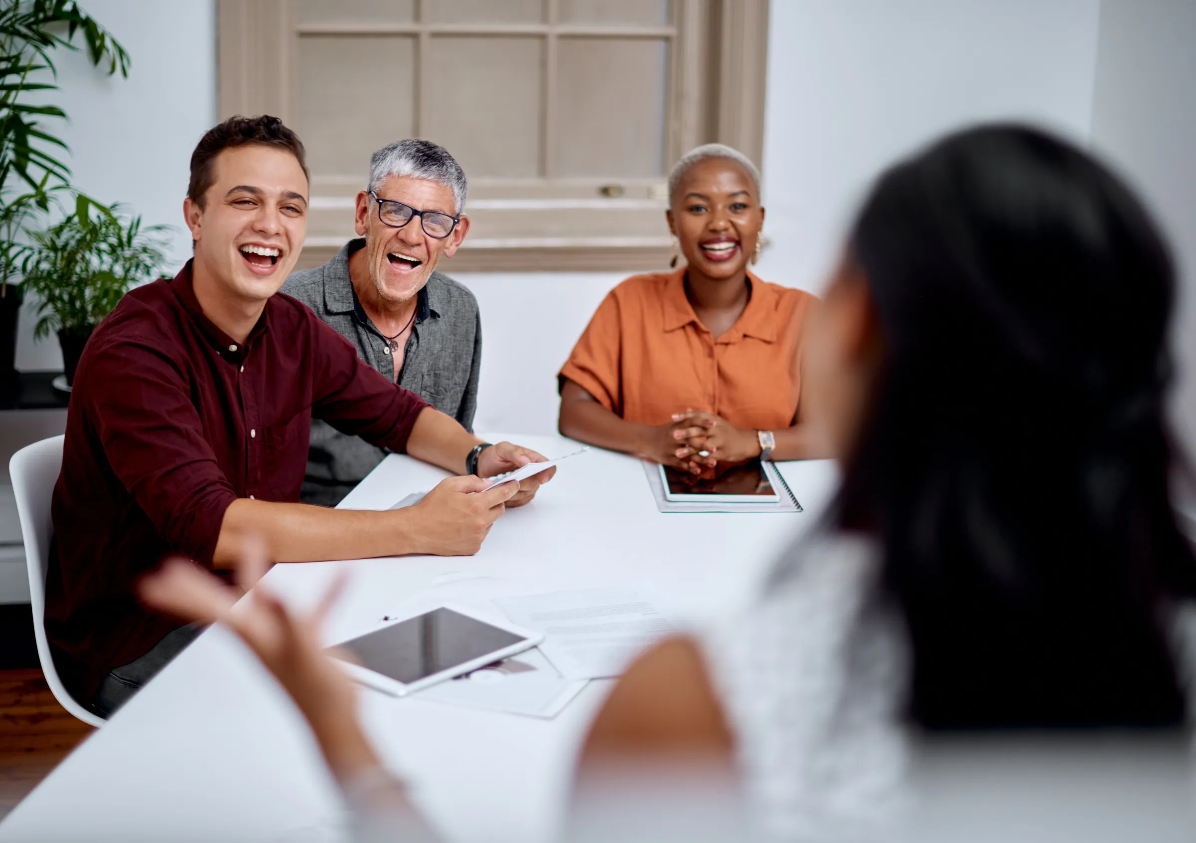 Three people smiling and engaged in a meeting with a person speaking, seated around a white table with tablets and documents.