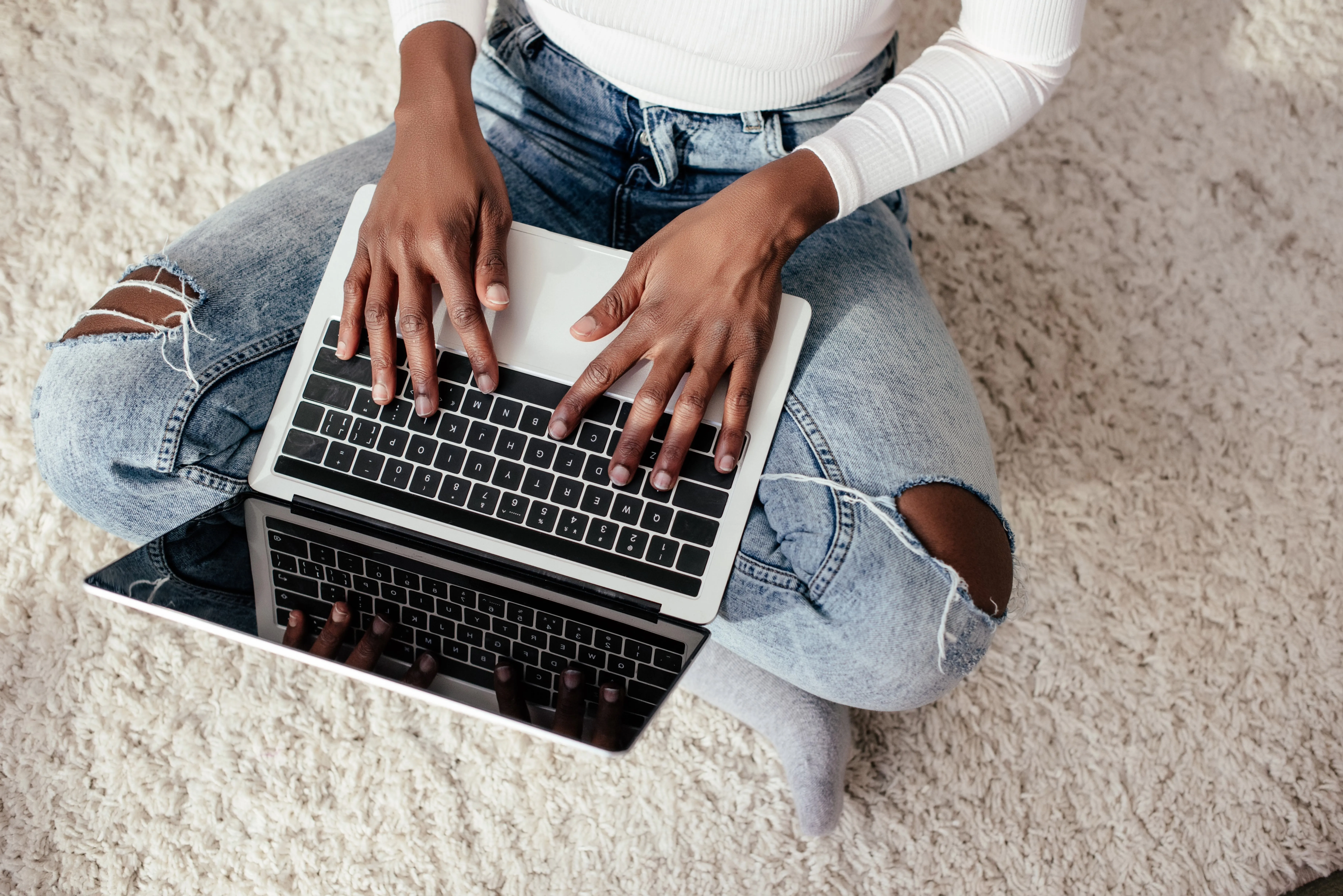 Person sitting cross-legged on a carpet typing on a laptop with ripped jeans and a white long-sleeve shirt.
