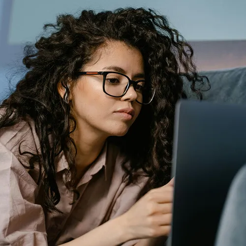 Young woman with curly hair and glasses wearing earbuds, looking at a laptop screen.