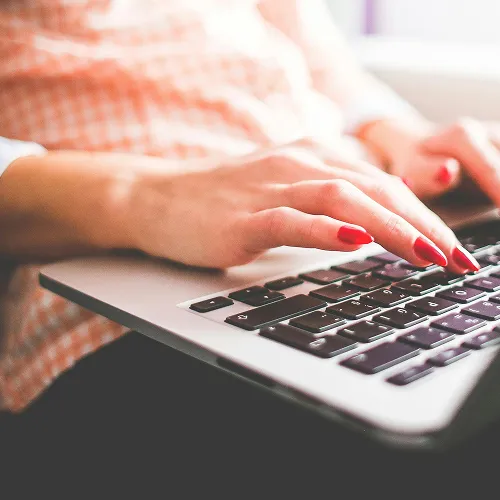 Close-up of a person typing on a laptop keyboard with red nail polish and wearing an orange checkered shirt.