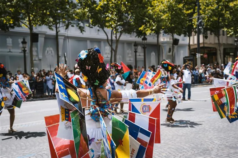Dancers in colorful costumes adorned with various national flags perform in an outdoor parade with spectators and trees in the background.
