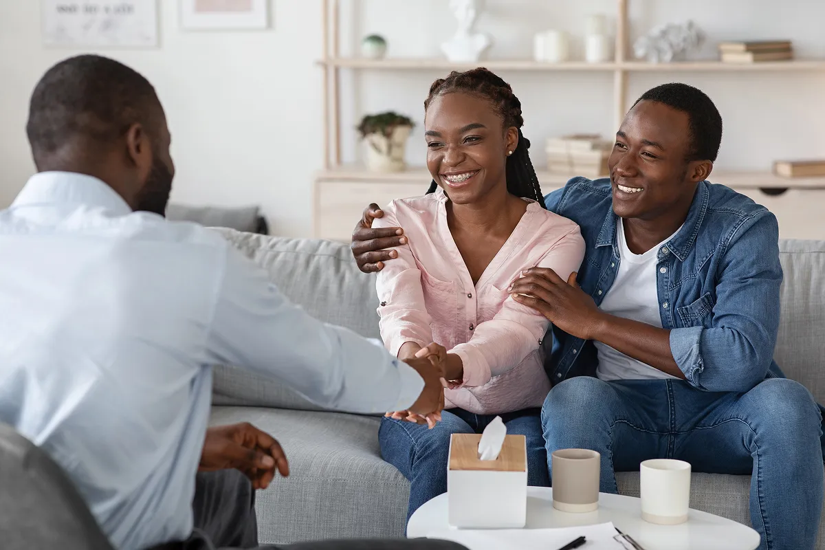 Smiling young couple sitting on a couch shaking hands with a man opposite them in a casual meeting setting.
