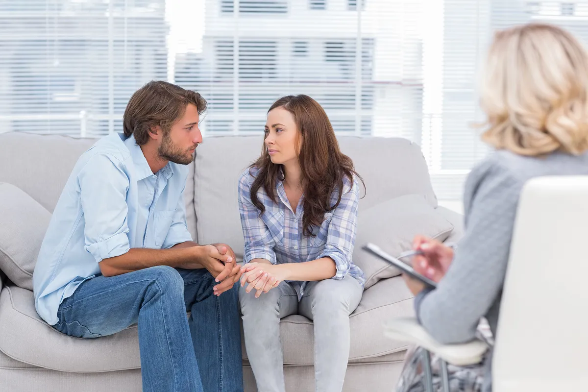 A couple seated on a couch attentively talking to a therapist who is taking notes.