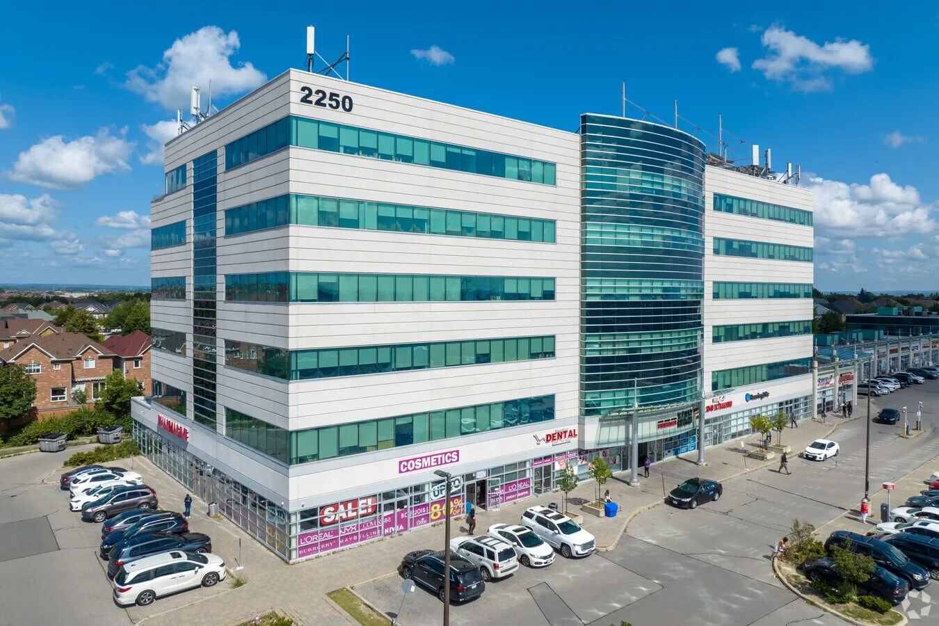 Five-story commercial office building with large glass windows, corner stores including a cosmetics shop with sale signs, and a parking lot with cars under a blue sky.