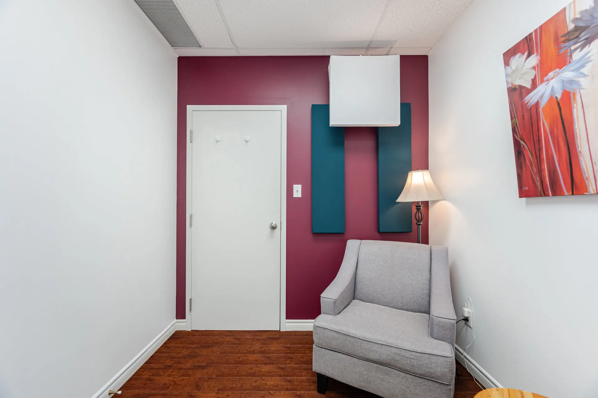 Small room with wooden floor, white and maroon walls, a gray armchair, a floor lamp, and a colorful floral painting.