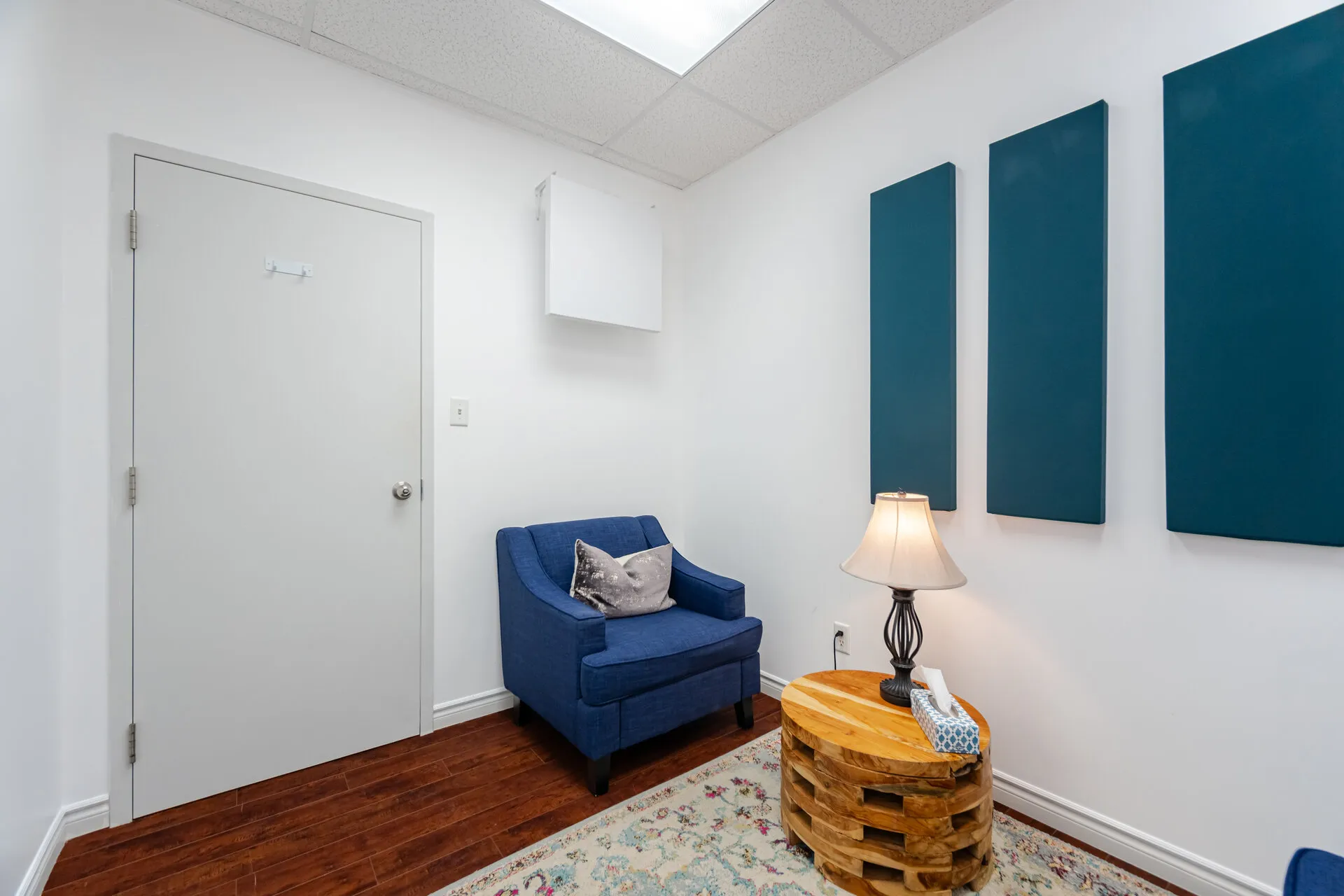 Cozy corner with a blue armchair, wooden round side table holding a lamp and tissues, three vertical blue wall panels, and hardwood floor with patterned rug.