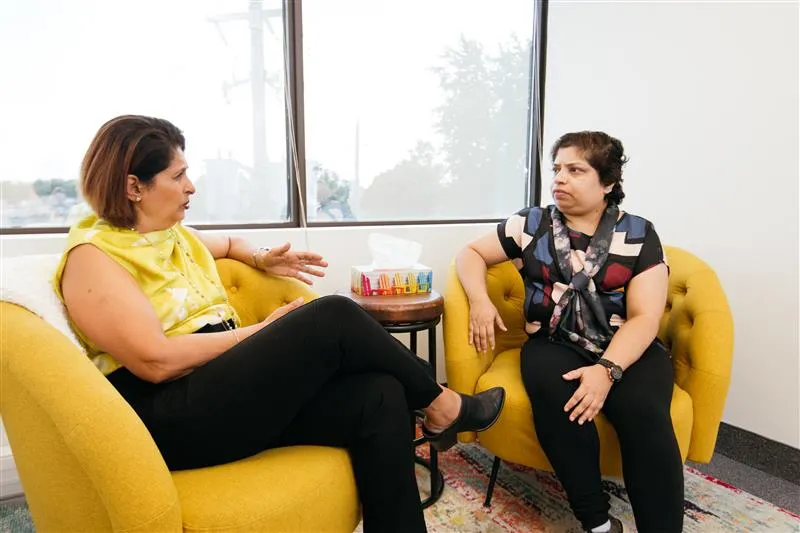 Two women seated in yellow armchairs engaged in a serious conversation in a bright room with a window in the background.