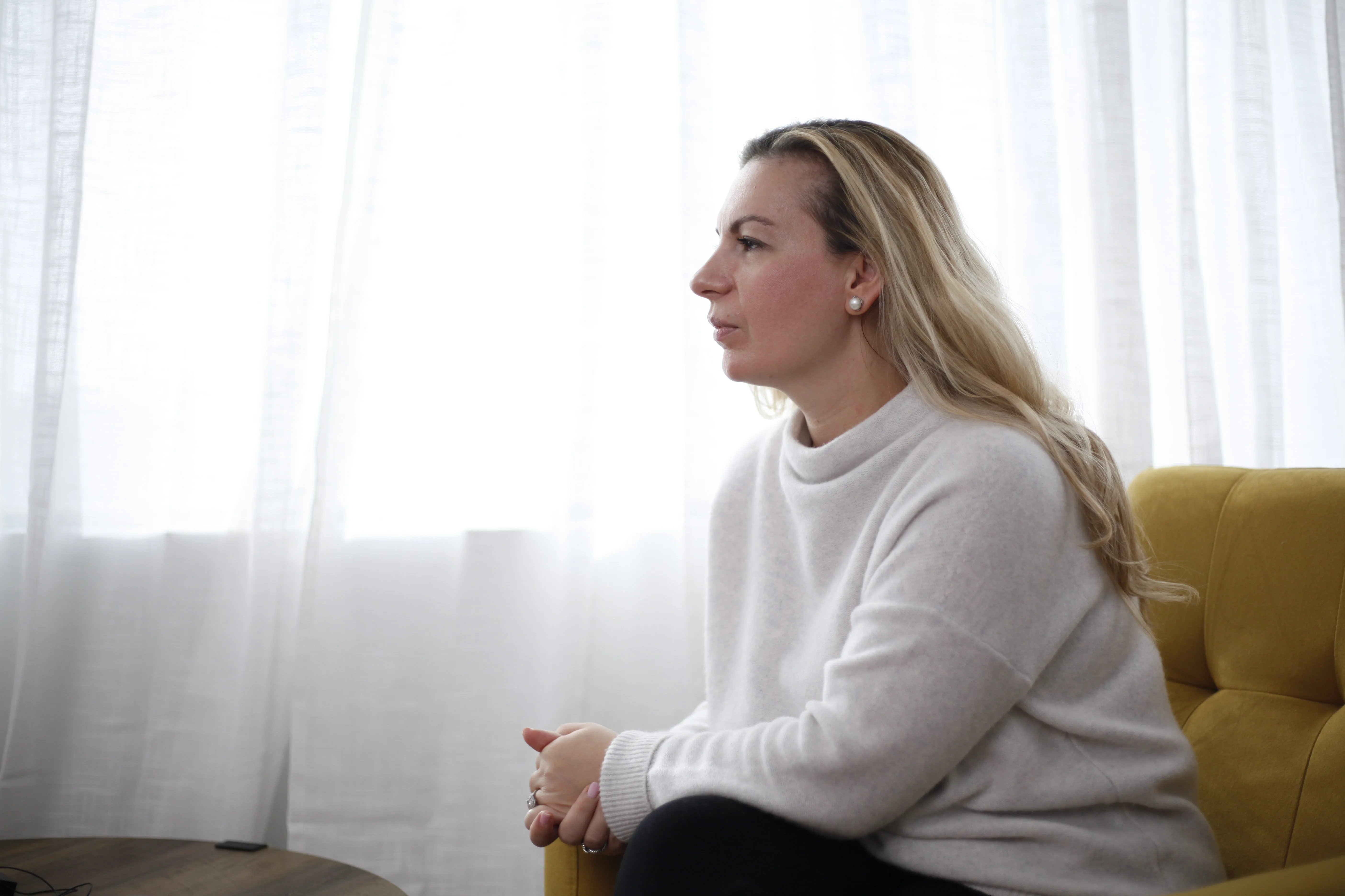 Woman with long blonde hair wearing a light gray sweater sitting on a yellow chair, looking thoughtfully to the right in front of white curtains.