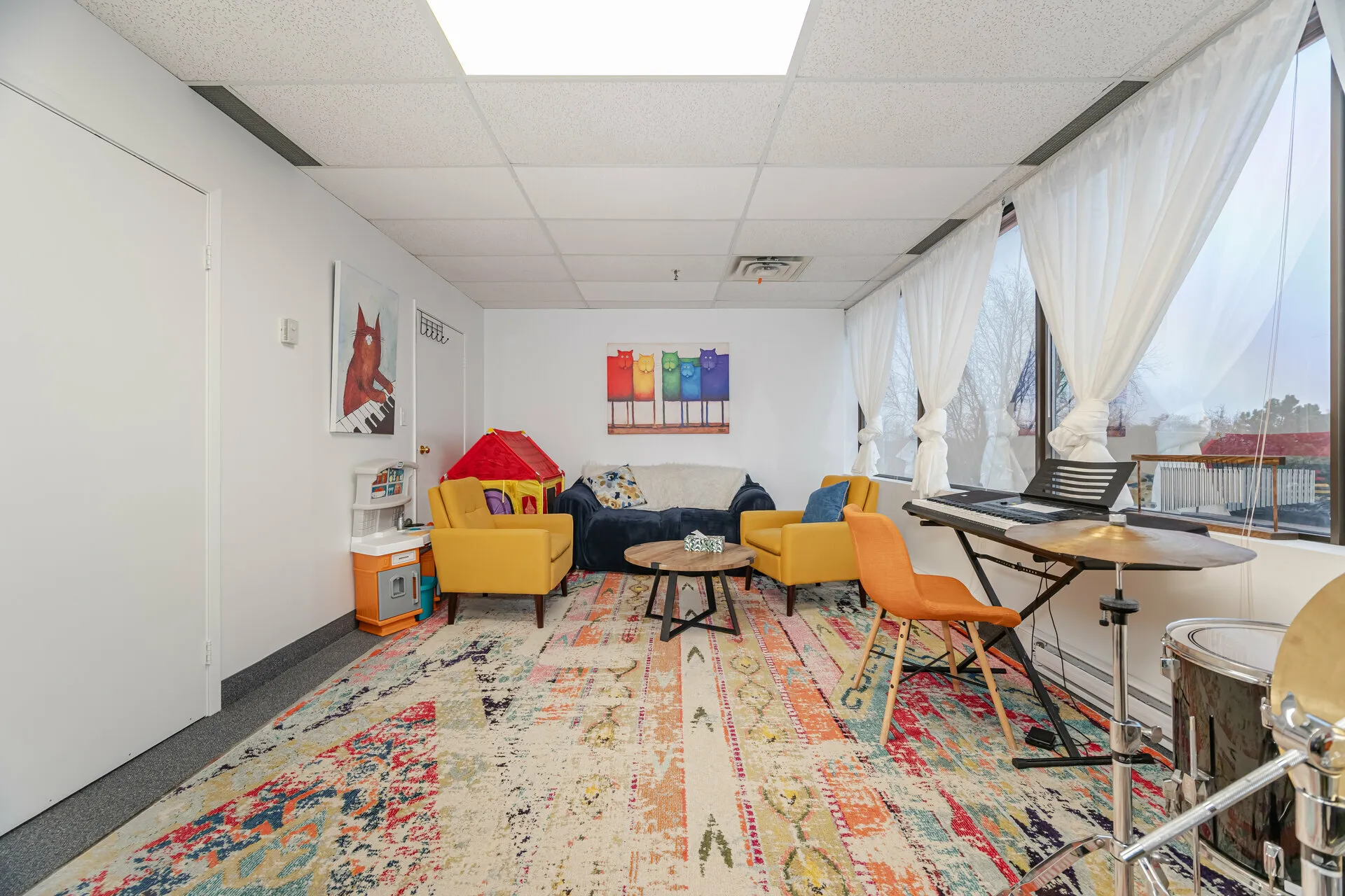 Bright room with colorful rug, yellow armchairs, blue couch, children's play kitchen and tent, musical keyboard, drum set, and large windows with white curtains.