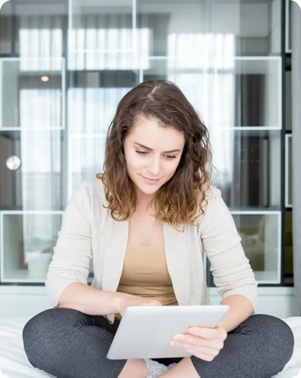 Woman with curly hair sitting cross-legged on a bed using a tablet device.
