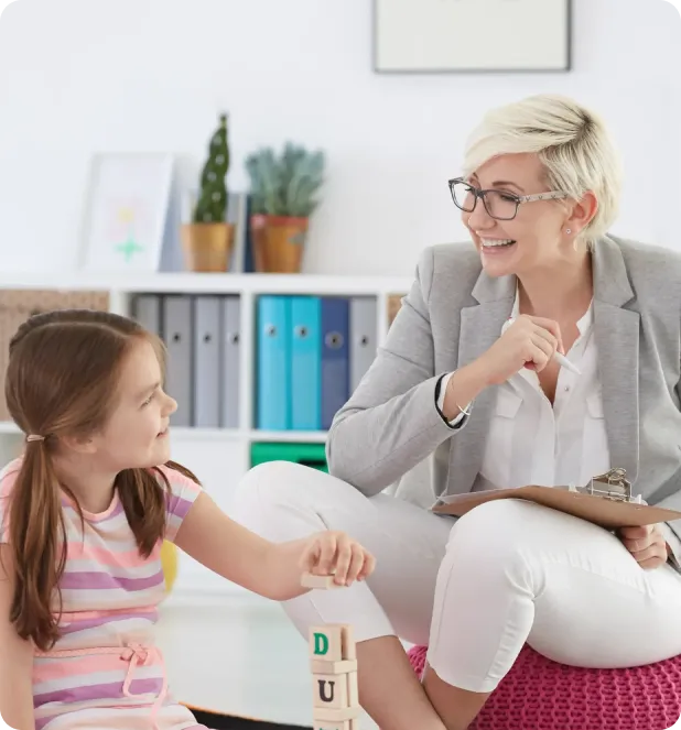 Smiling female therapist holding a clipboard interacts with a young girl playing with wooden blocks in a bright office.