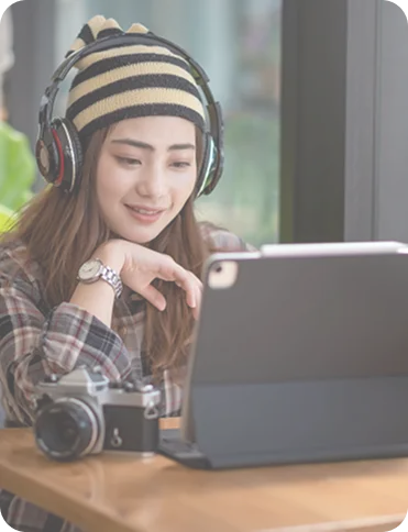Young woman wearing headphones and a striped beanie, smiling while using a tablet at a wooden table with a camera nearby.