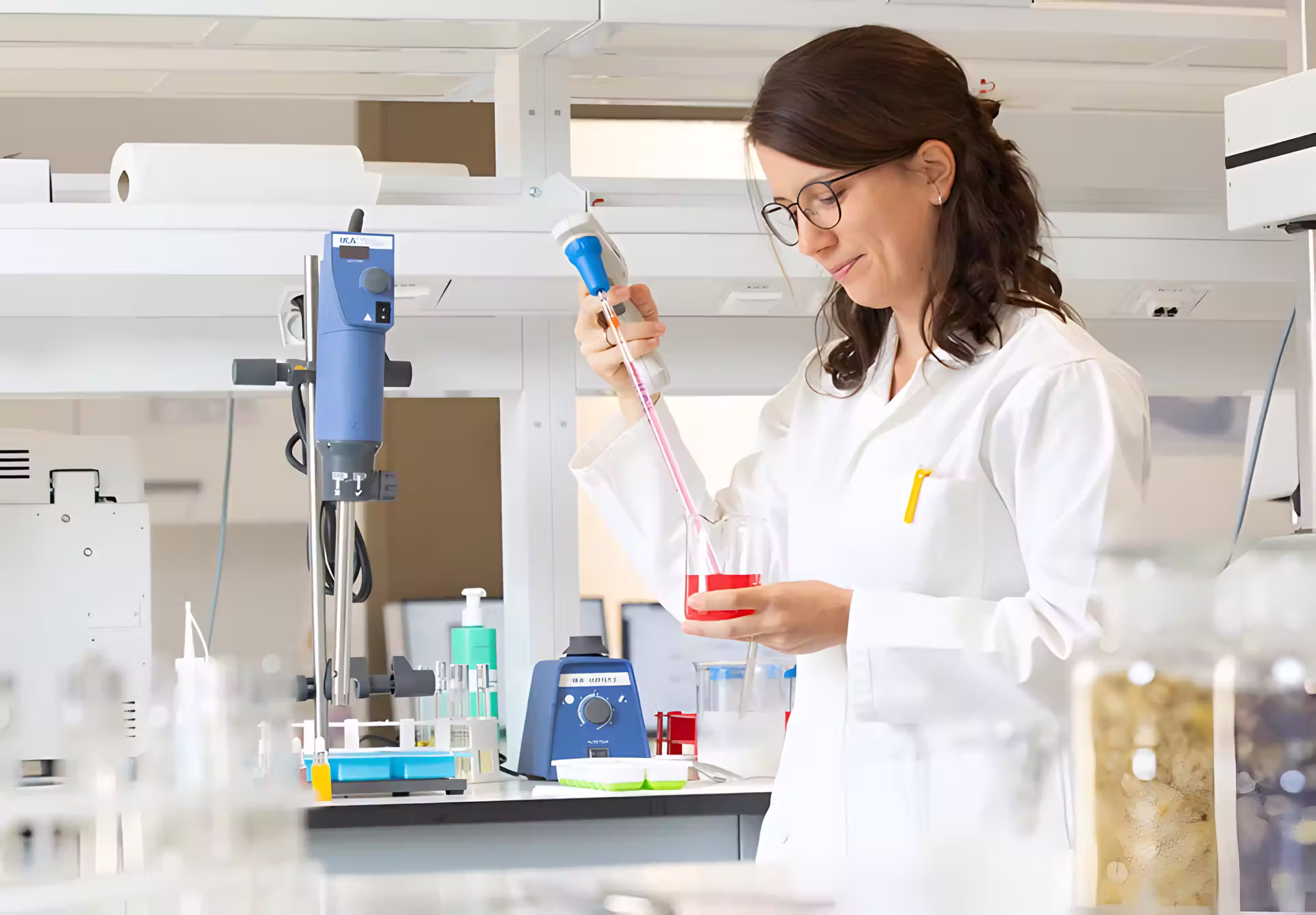 Weibliche Wissenschaftlerin in weißem Laborkittel und Brille führt Forschung mit Laborausrüstung und farbigen Lösungen in professionellem Labor durch