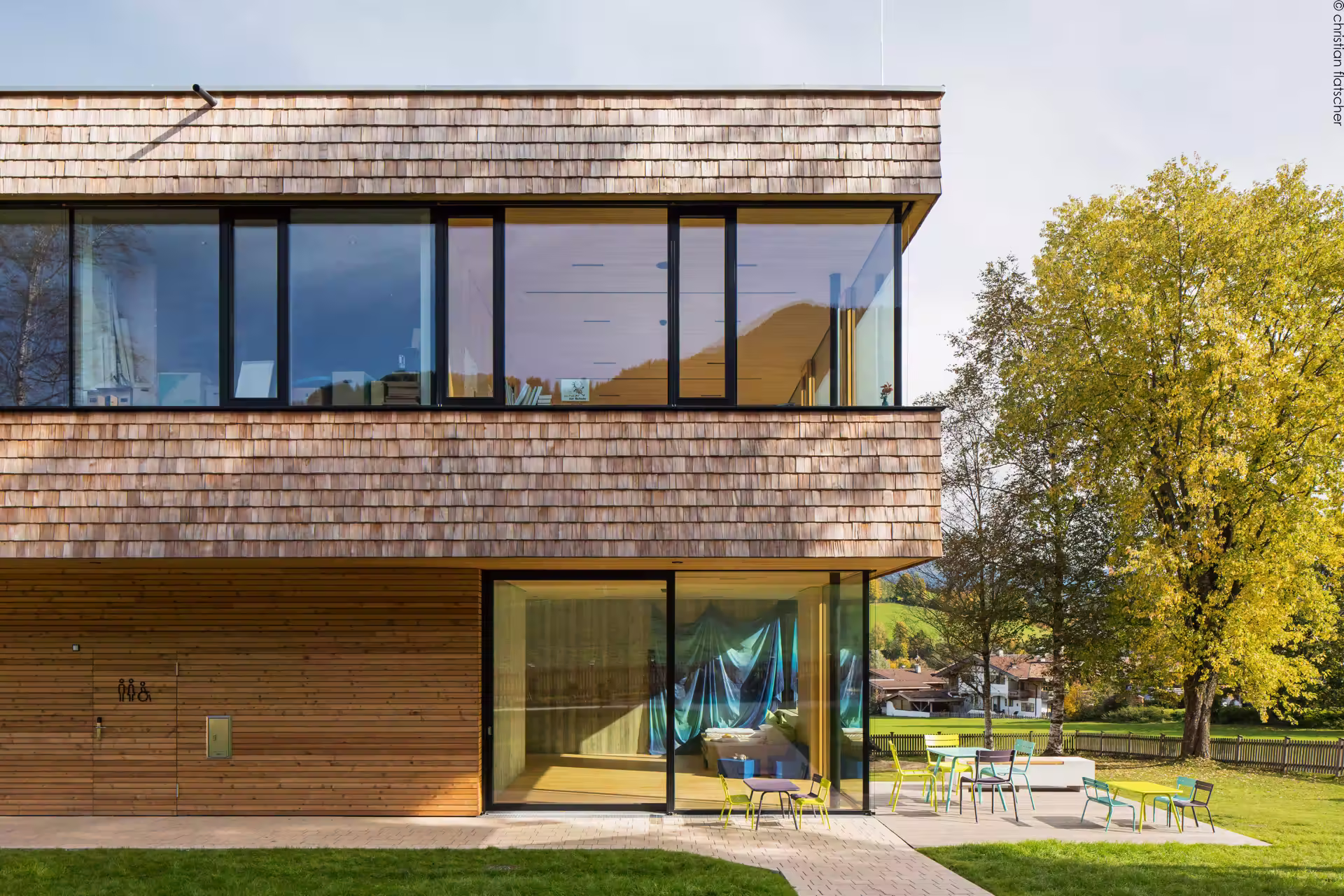 Modernes Holzhaus mit Schindelverkleidung, großen Glasfenstern im Obergeschoss, horizontaler Holzverkleidung im Erdgeschoss, umgeben von grünem Rasen und Bäumen