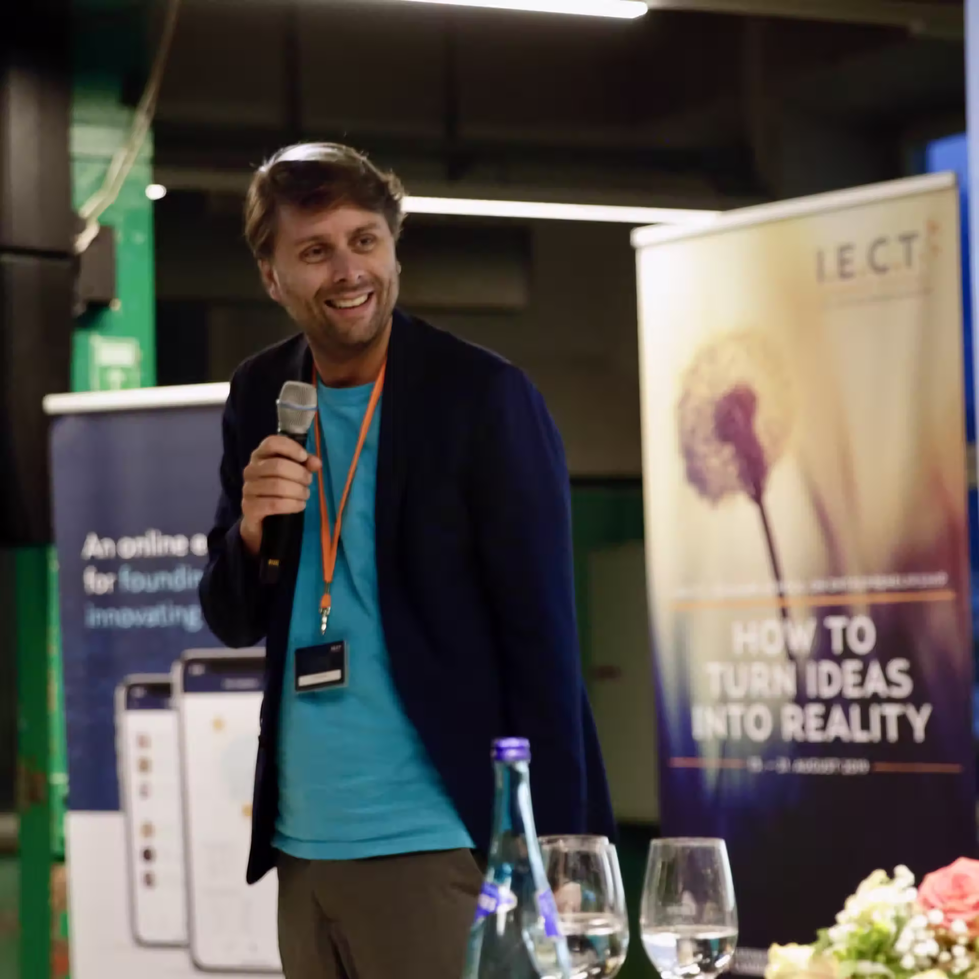 Enthusiastischer Sprecher in marineblauen Blazer und türkisfarbenem Hemd bei I.E.C.T.-Veranstaltung, hält Mikrofon mit Konferenz-Bannern 'Turn Ideas Into Reality' im Hintergrund