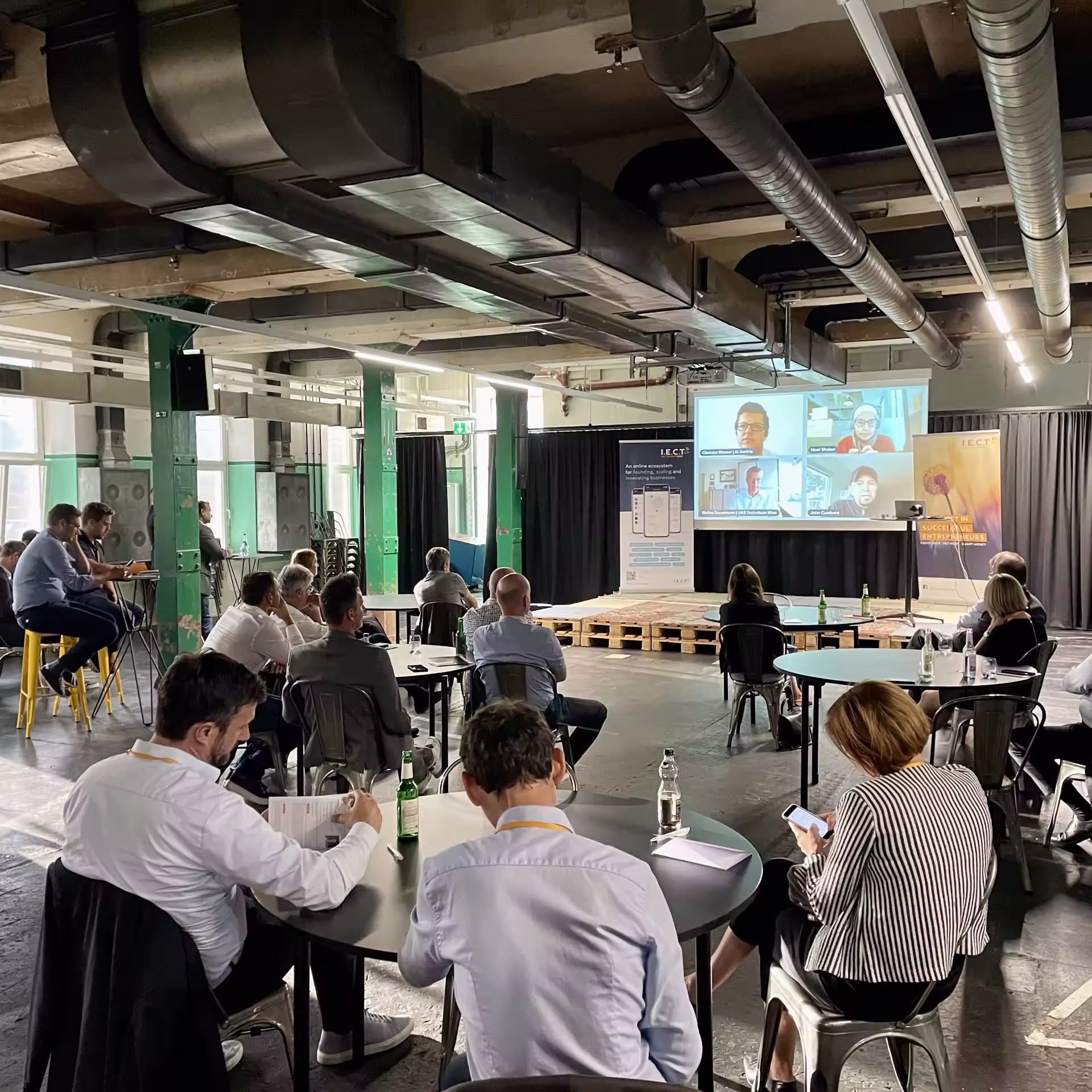 Moderner Konferenzraum mit sichtbarer Industriedecke, grünen Struktursäulen und Teilnehmern an runden Tischen, die eine Präsentation mit Videokonferenz-Teilnehmern auf dem Bildschirm verfolgen