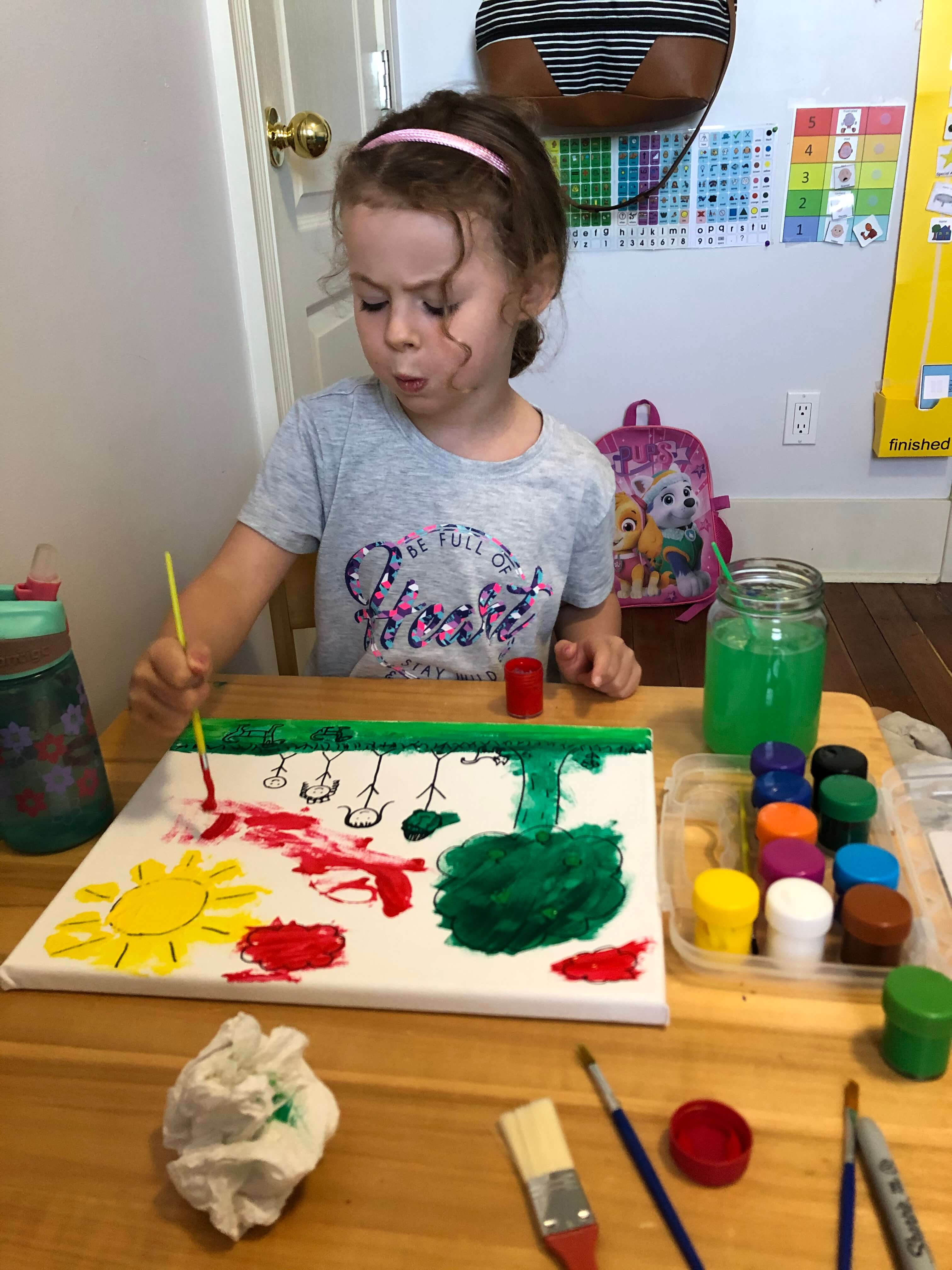 Young girl painting a colorful picture with a sun, tree, and figures at a wooden table.