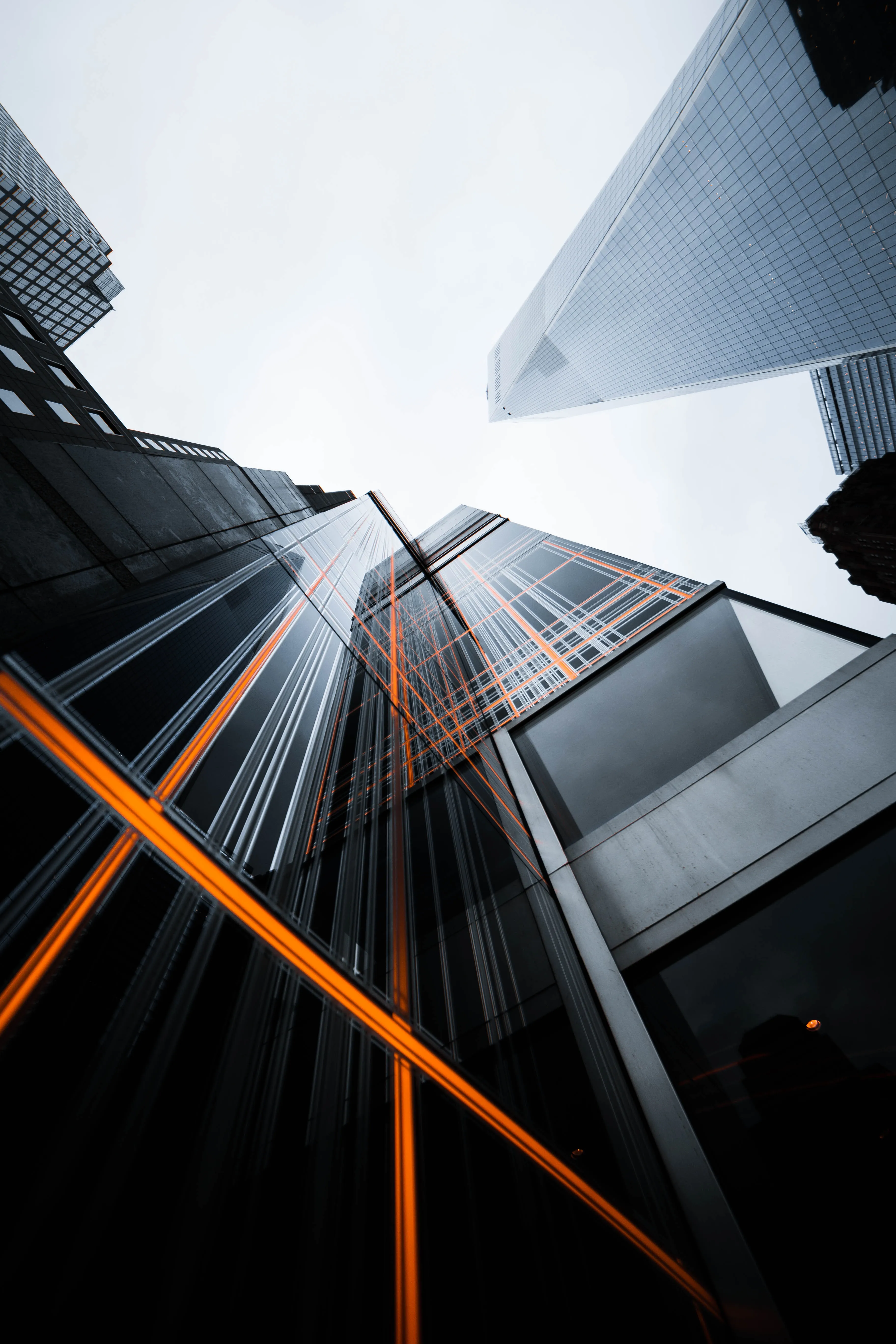 Low-angle view of modern skyscrapers with reflective glass and orange structural highlights against a gray sky.