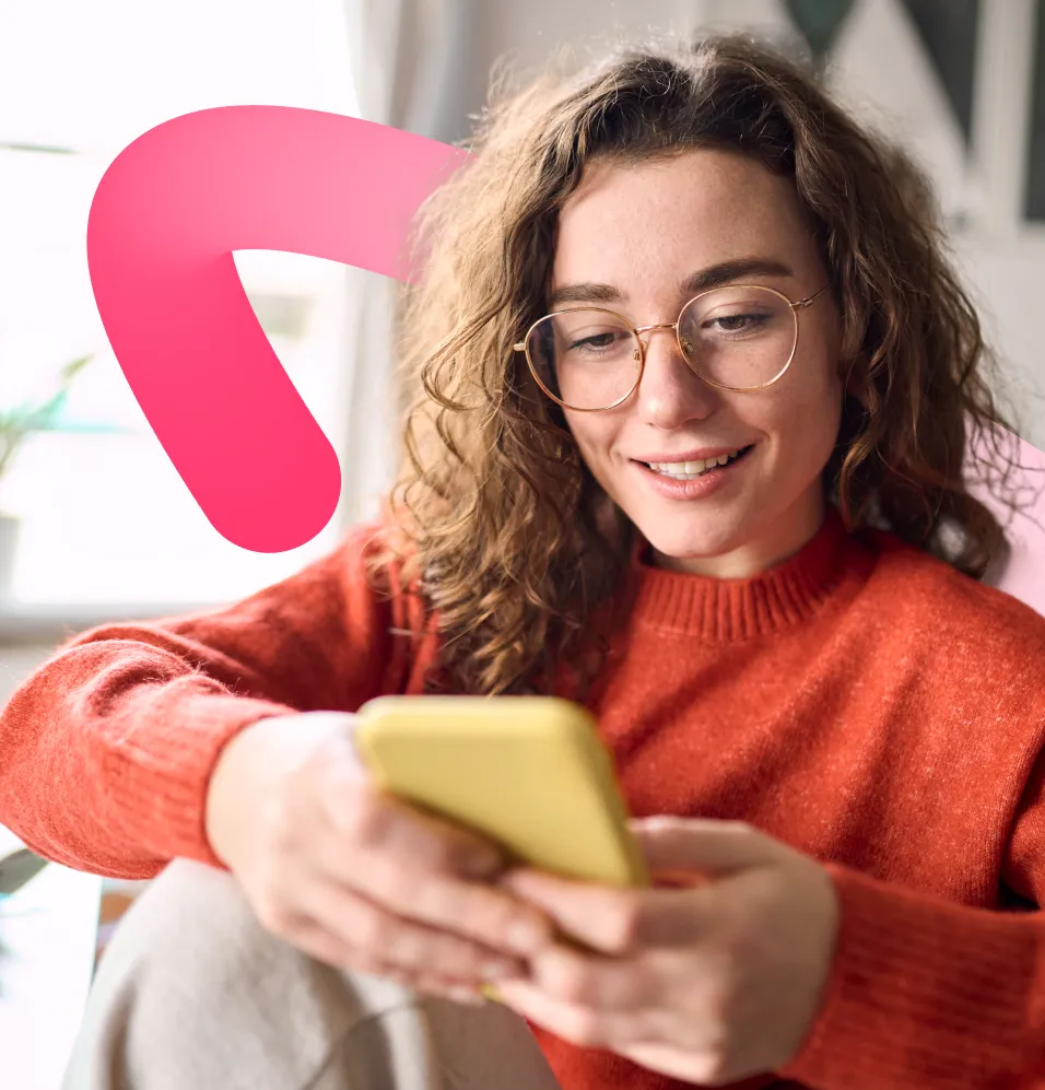 Woman with curly hair and glasses smiling while using a yellow smartphone indoors.