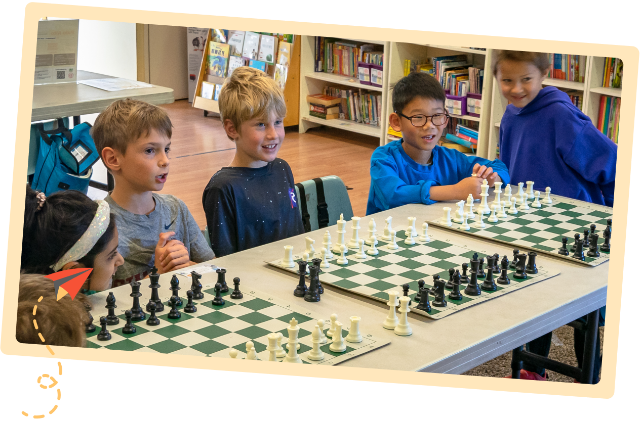 A group of engaged children smile and interact around chess boards during a class at the Palo Alto