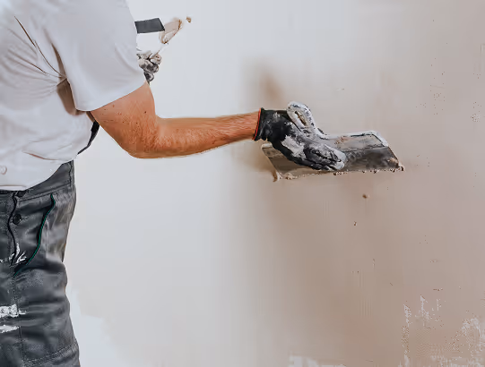 Handwerker trägt weiße Kleidung und schwarze Handschuhe und trägt Spachtelmasse an eine Wand auf.