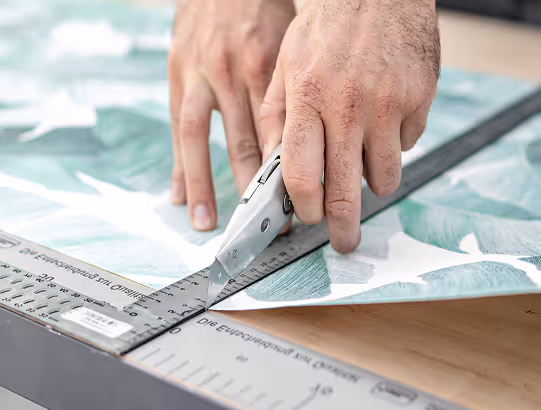 Hände schneiden Tapete entlang eines Metalllineals mit einem Cutter-Messer.